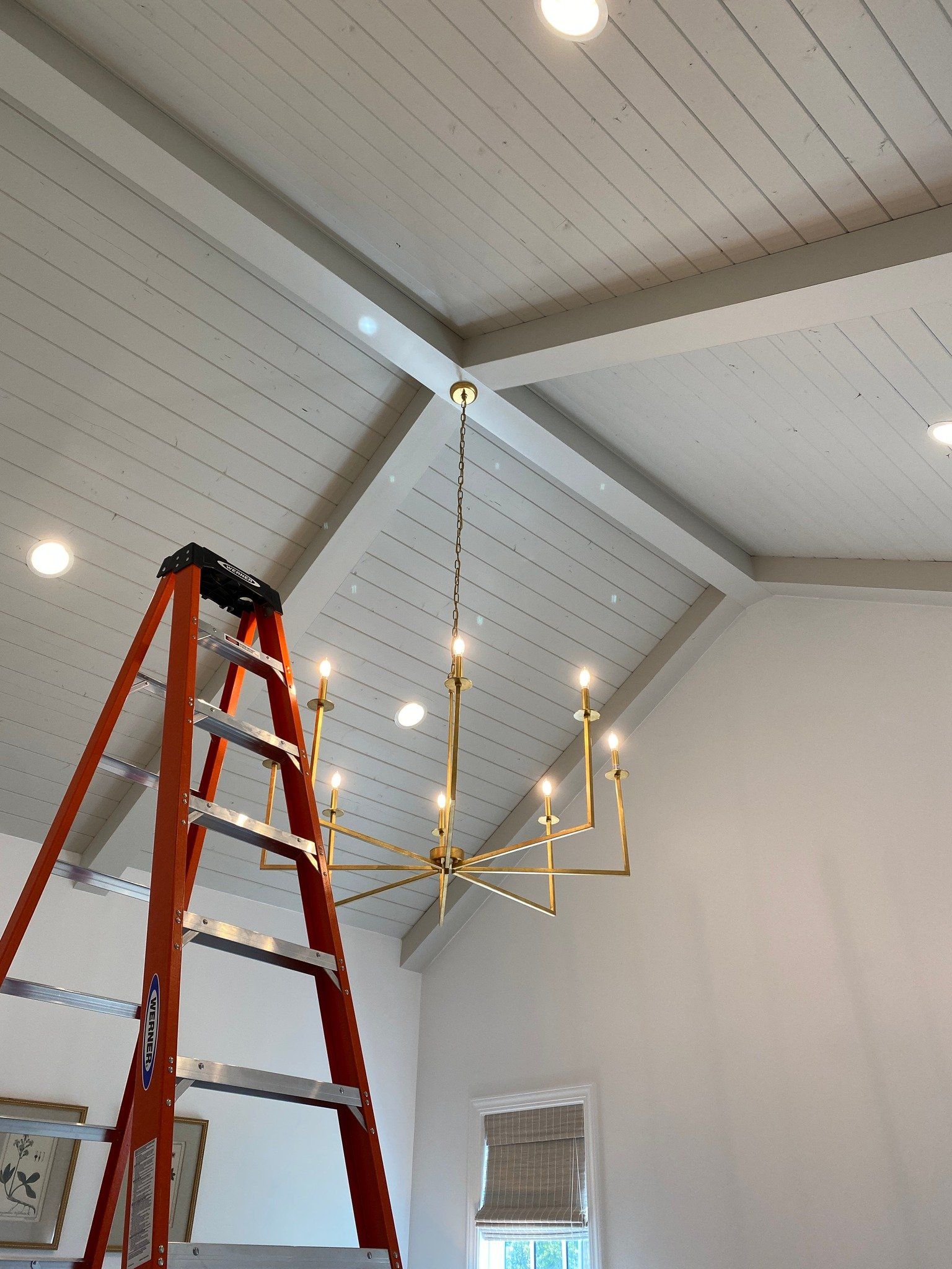 Orange ladder beneath a gold chandelier hanging from a white sloped ceiling with recessed lighting.