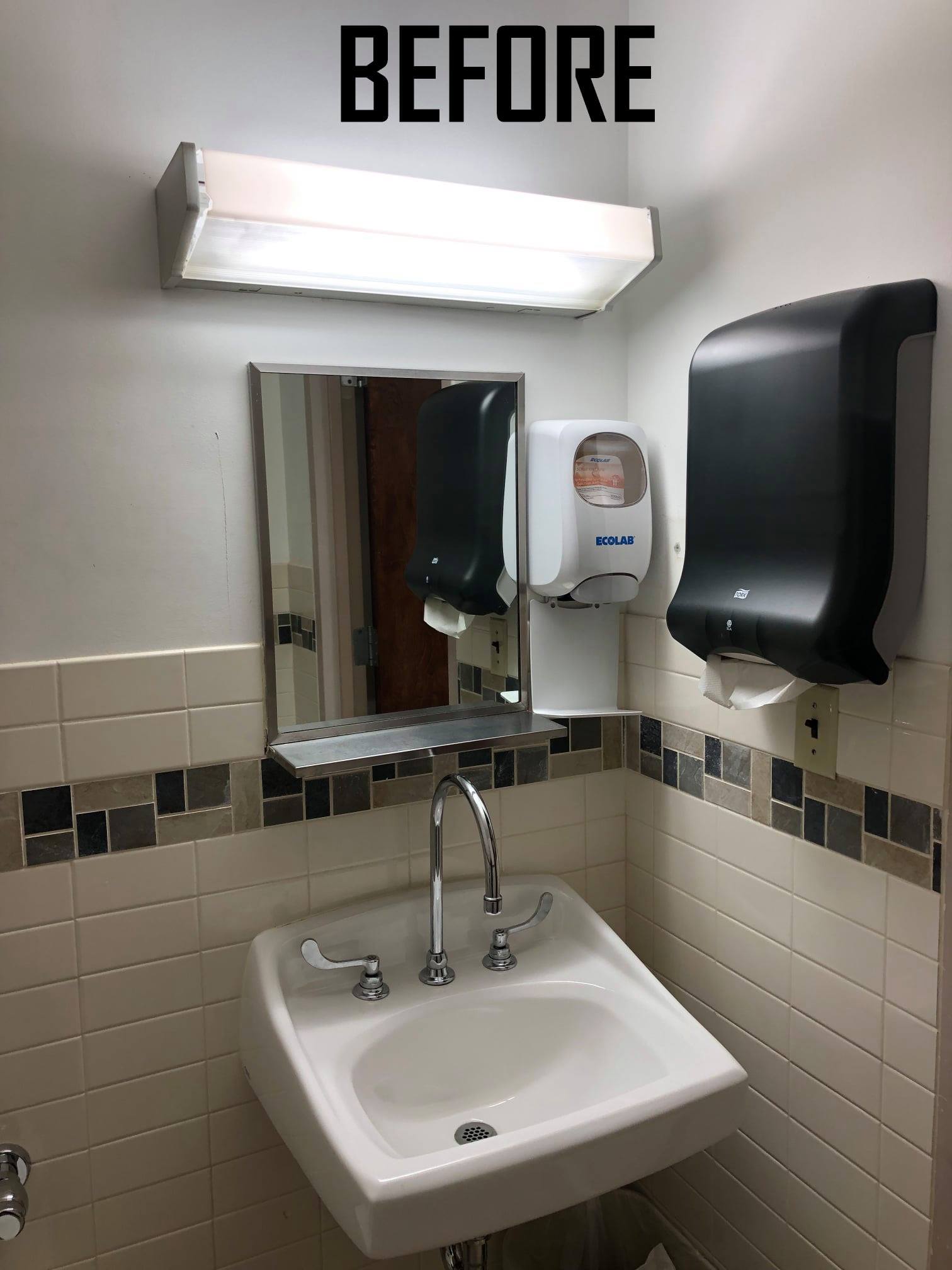 A public restroom sink with mirror, dispensers, and overhead fluorescent light. Beige and brown tile.