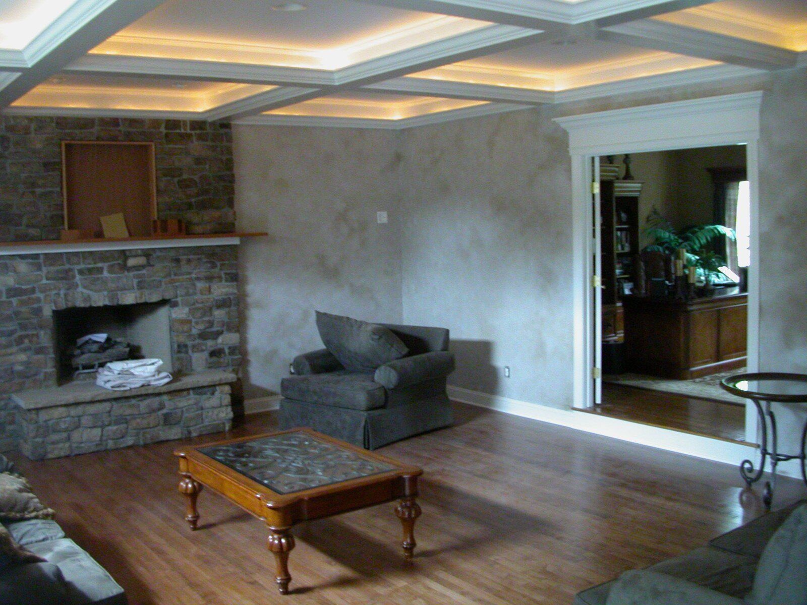 Living room with stone fireplace, wood floors, and coffered ceiling with ambient lighting.