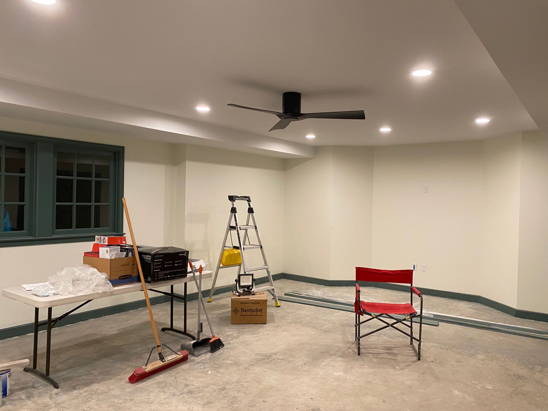 A room under construction with a ceiling fan, ladder, table, and chair. Walls are painted off-white.