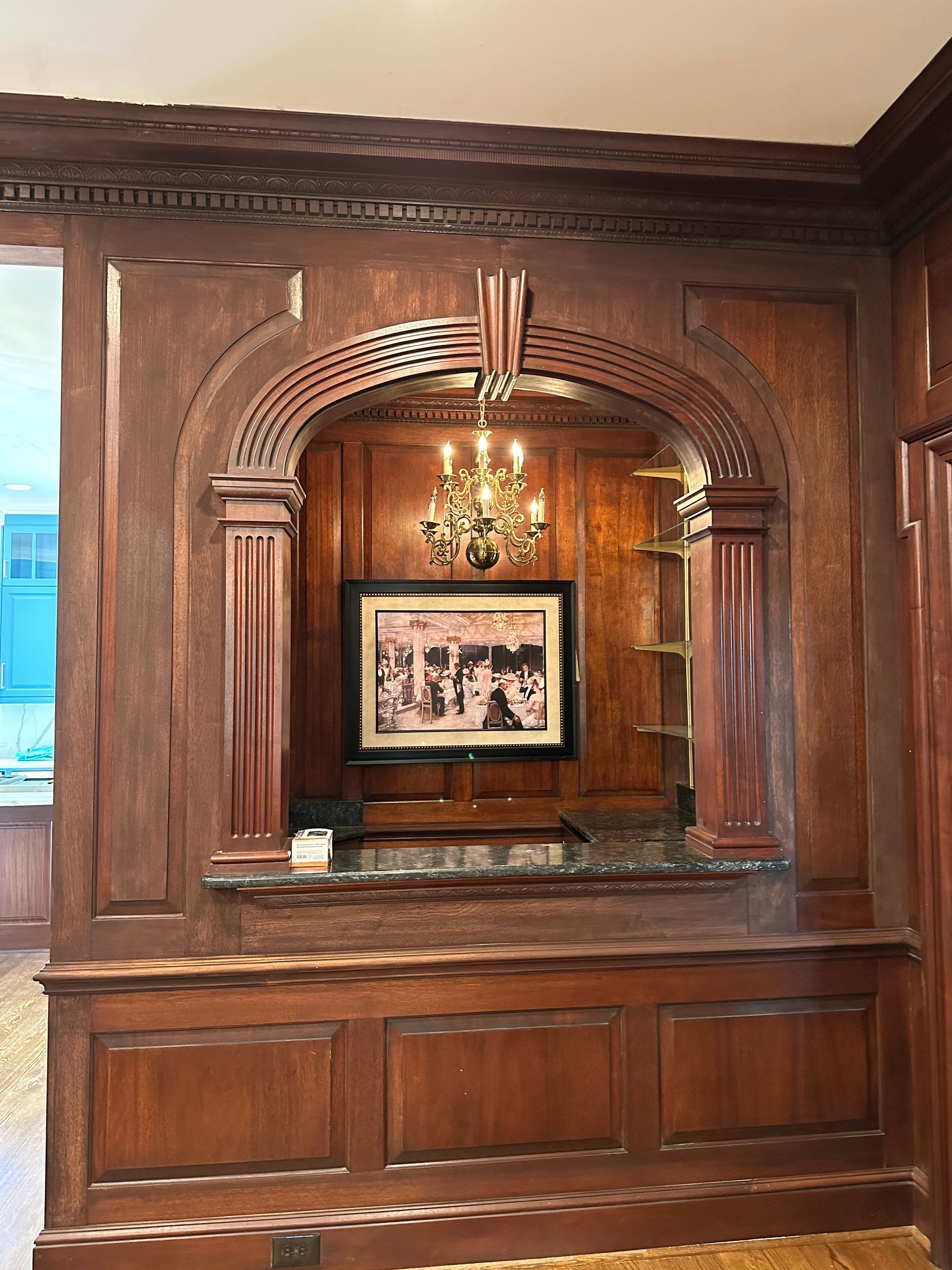 Dark wood paneled wall with an arched alcove displaying a framed photo, chandelier, and built-in shelves.