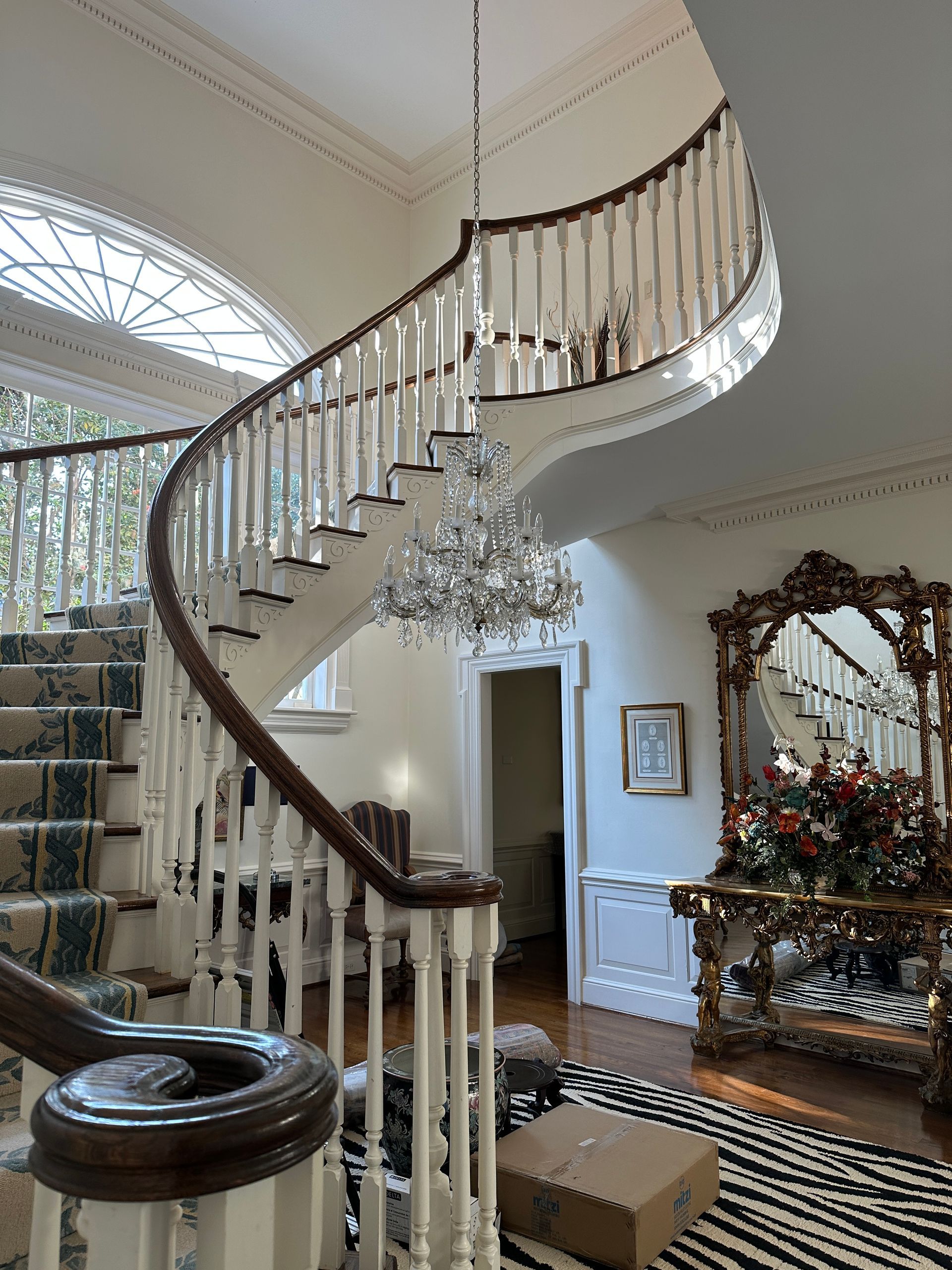 Elegant spiral staircase with crystal chandelier, decorative mirror, and patterned rug.