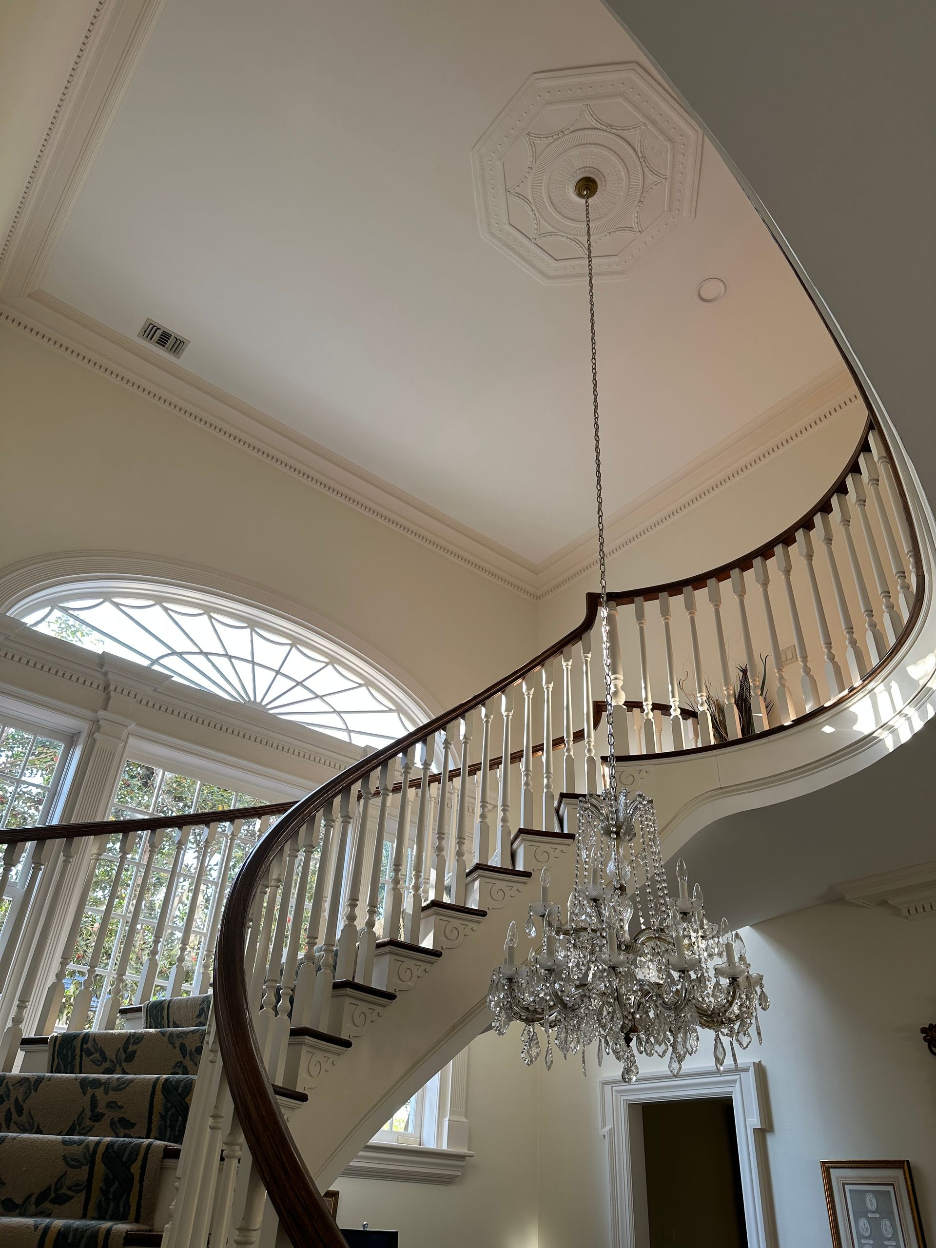 Elegant curved staircase with chandelier, white walls, and a large arched window.