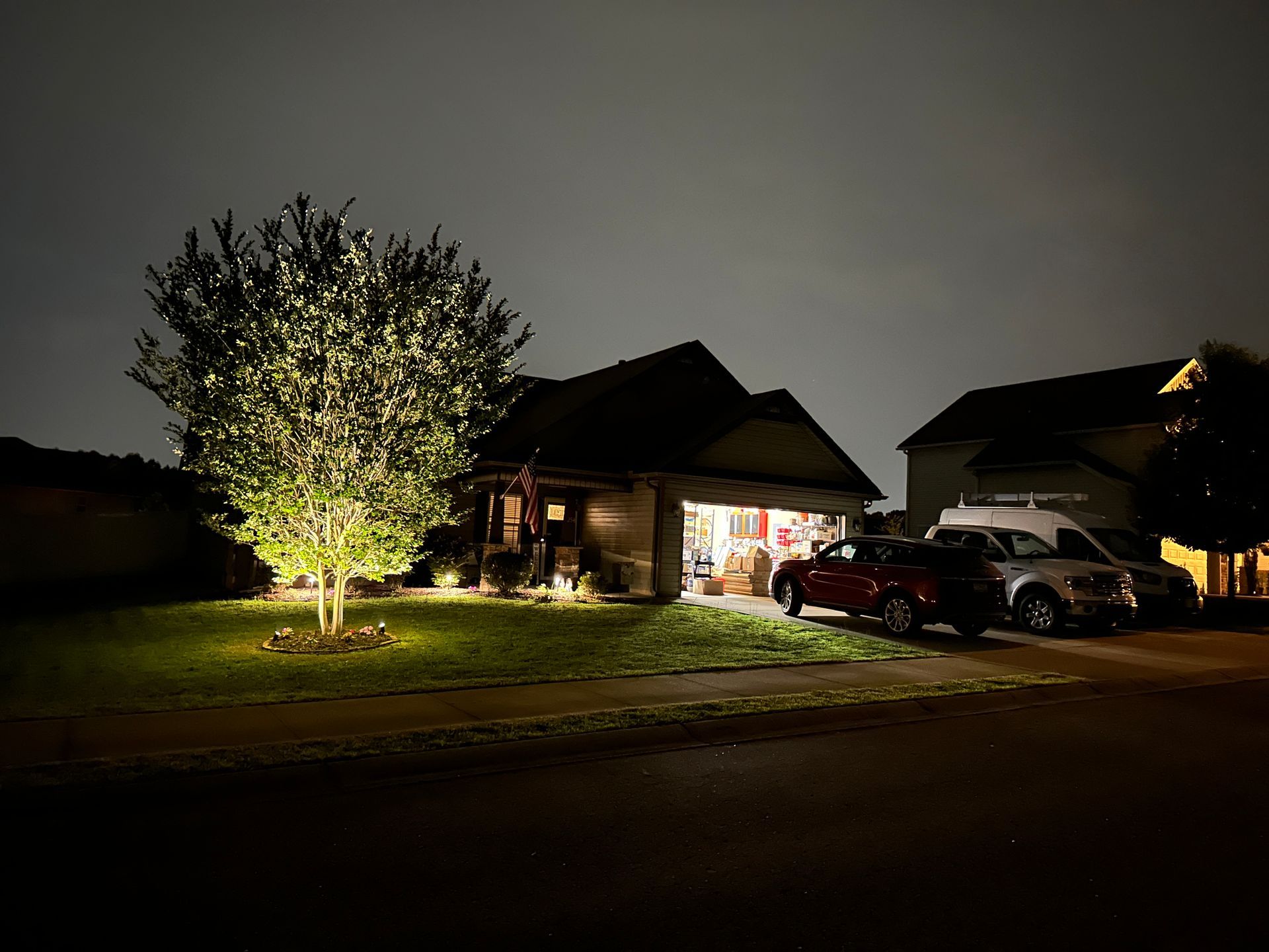 Nighttime view of a house with illuminated tree and garage; cars parked in driveway.