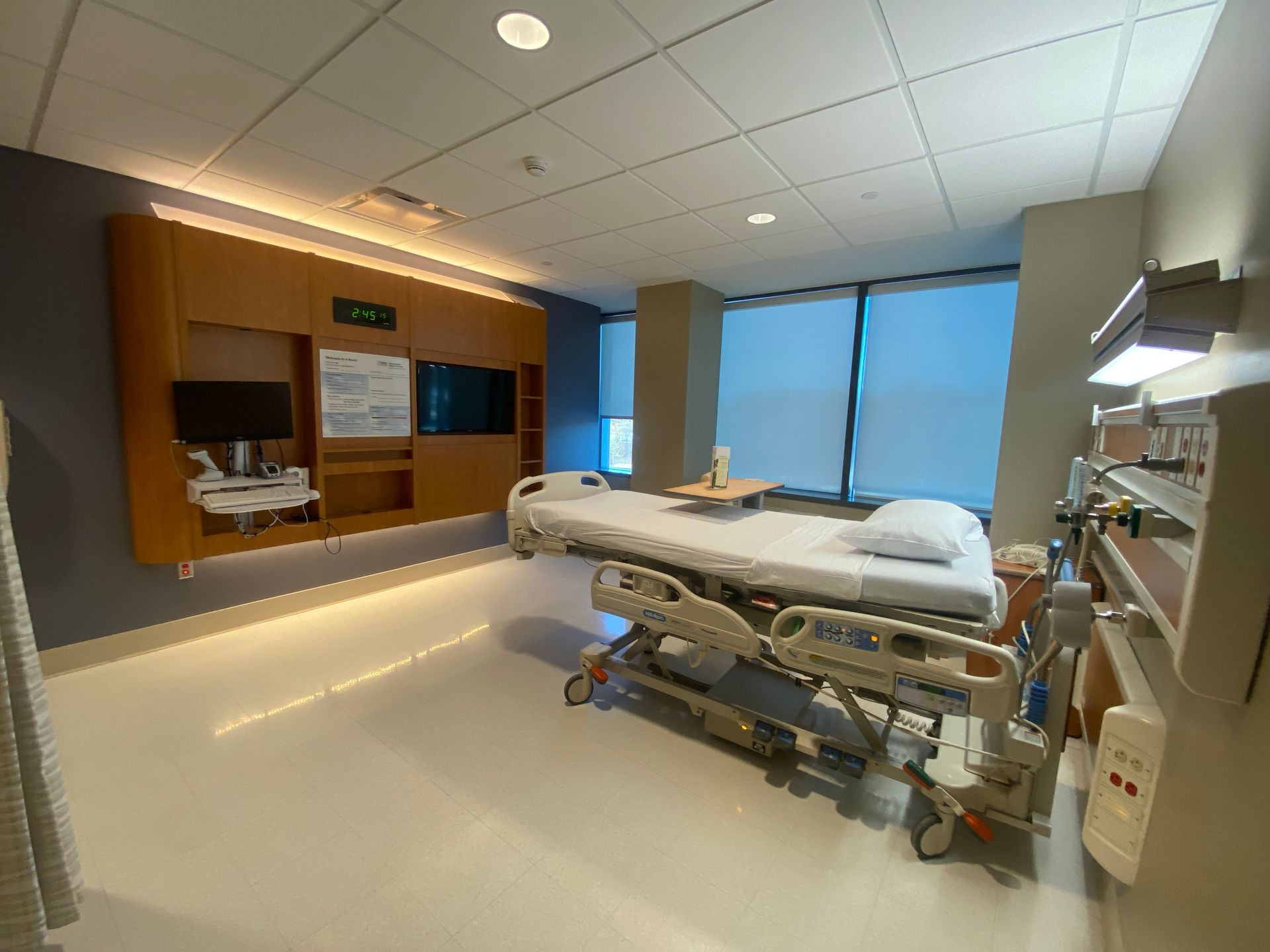 Hospital room with a bed, medical equipment, a window, and wood cabinetry on the wall.