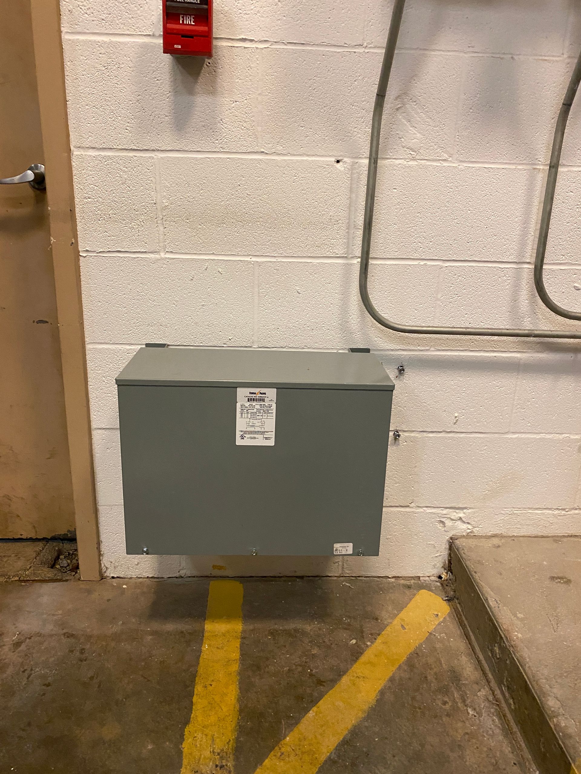 Gray electrical box mounted on a concrete block wall near yellow lines on floor, next to a door.