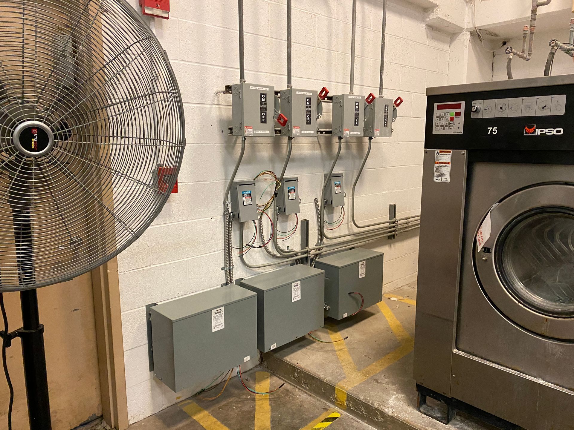 Industrial laundry room with machinery and electrical equipment. Large fan, washing machine, and control boxes on the wall.