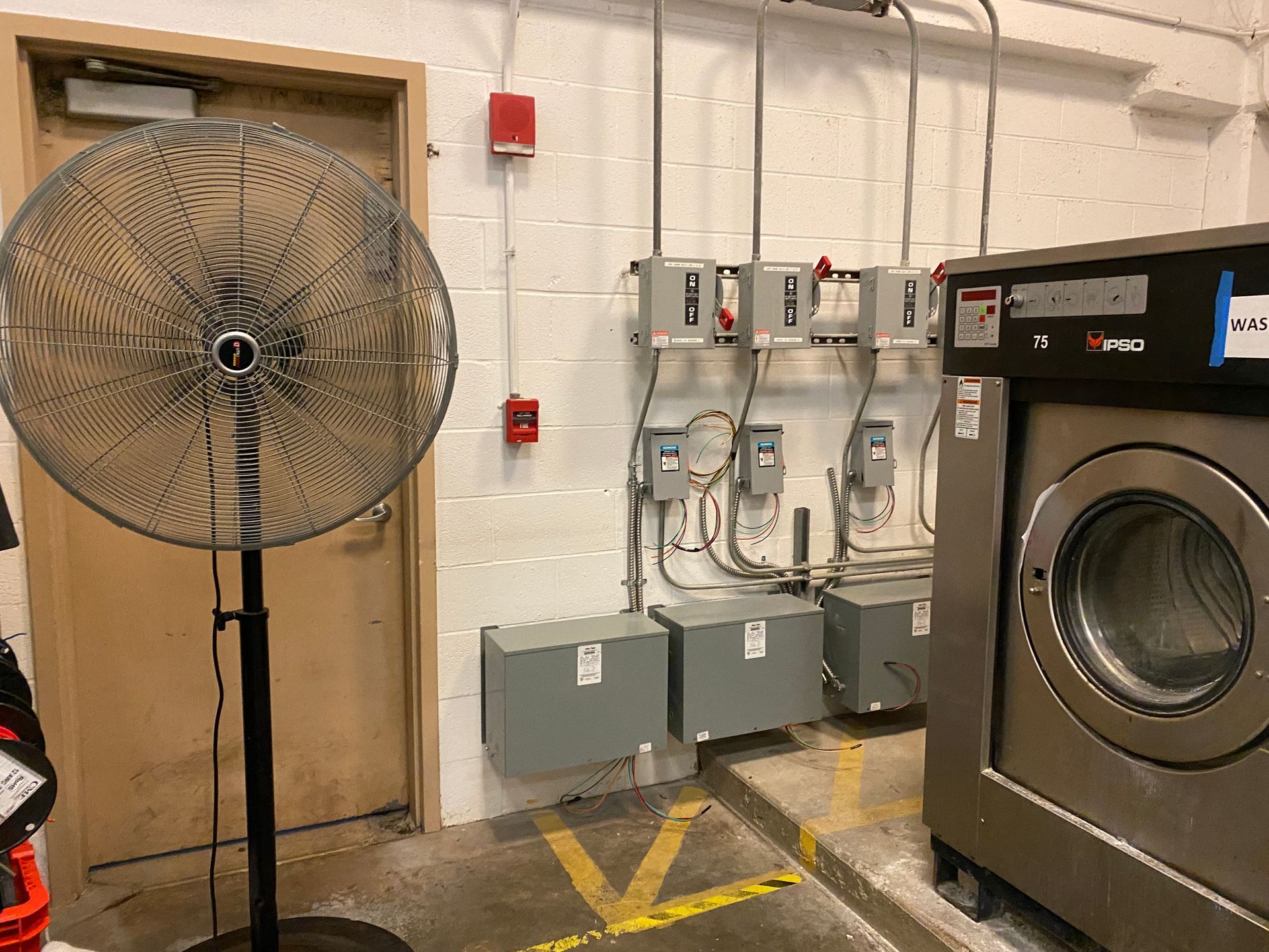 Industrial laundry room with a standing fan, washing machine, and electrical boxes.