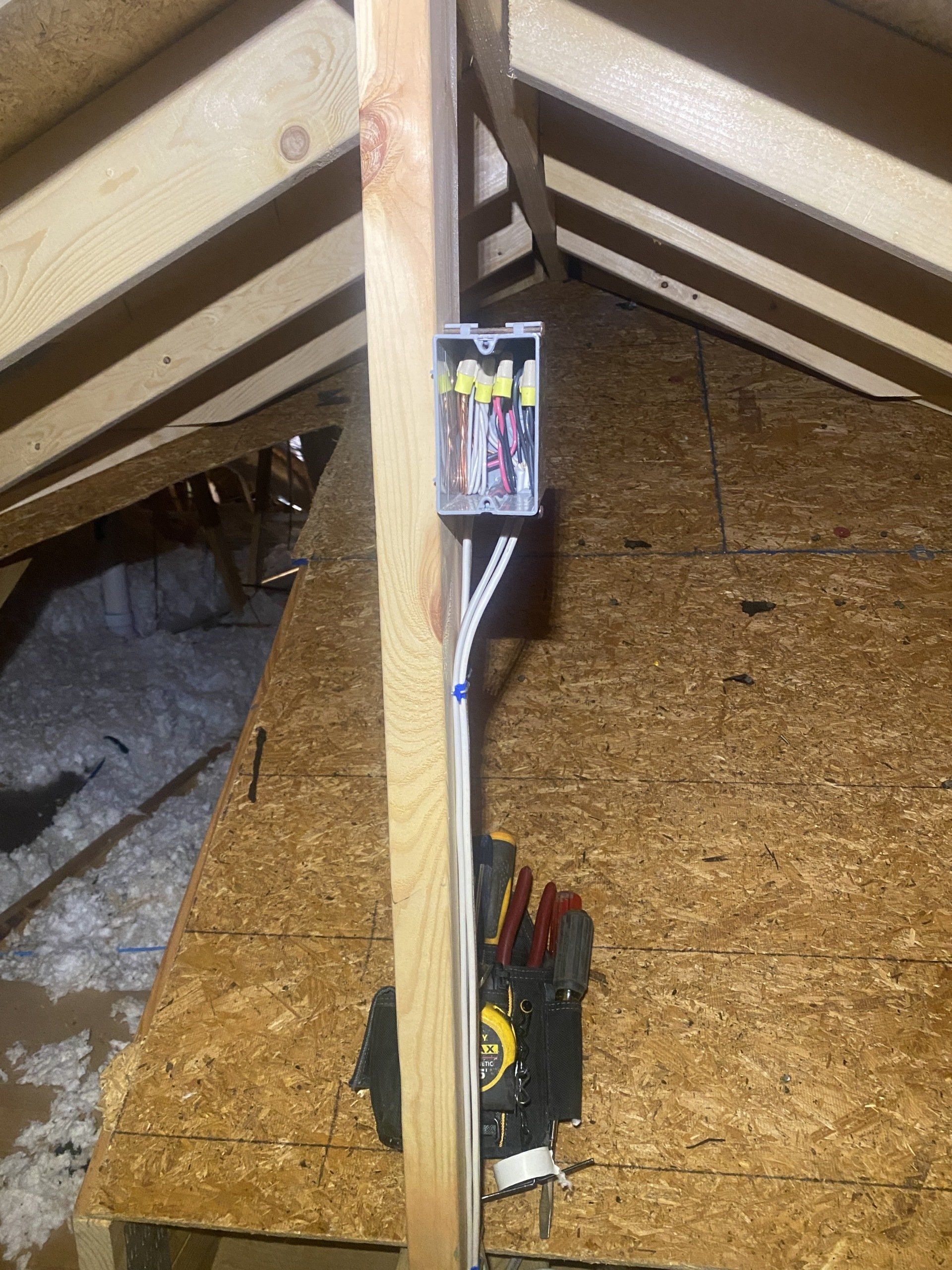 Electrical junction box mounted on a wooden beam in an attic. White wires are visible, along with tools below.