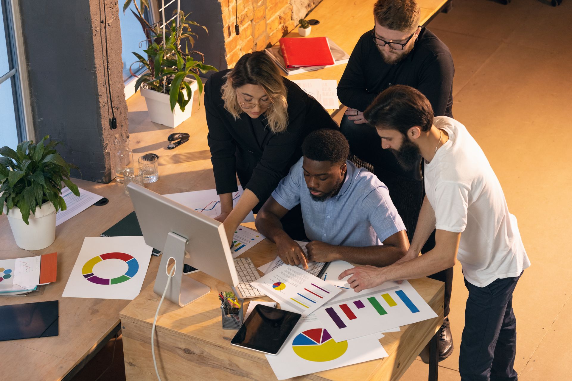 A diverse group of colleagues reviews colorful data charts and graphs together around a wooden desk in an office.