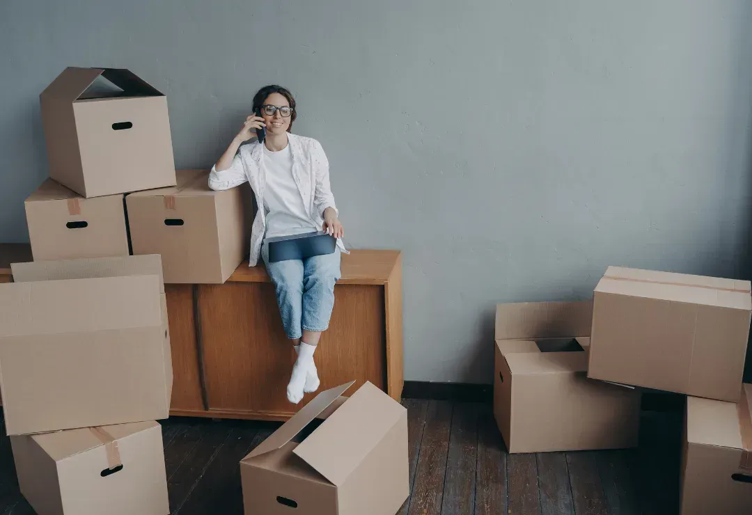 a woman sitting on a desk on the phone next to a bunch of cardboard boxes