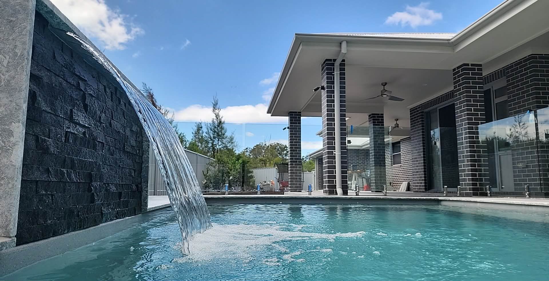Pool with waterfall feature; modern home exterior in background. Blue water and sky. — Clearwater Pools Hallidays Point In Hallidays Point, NSW