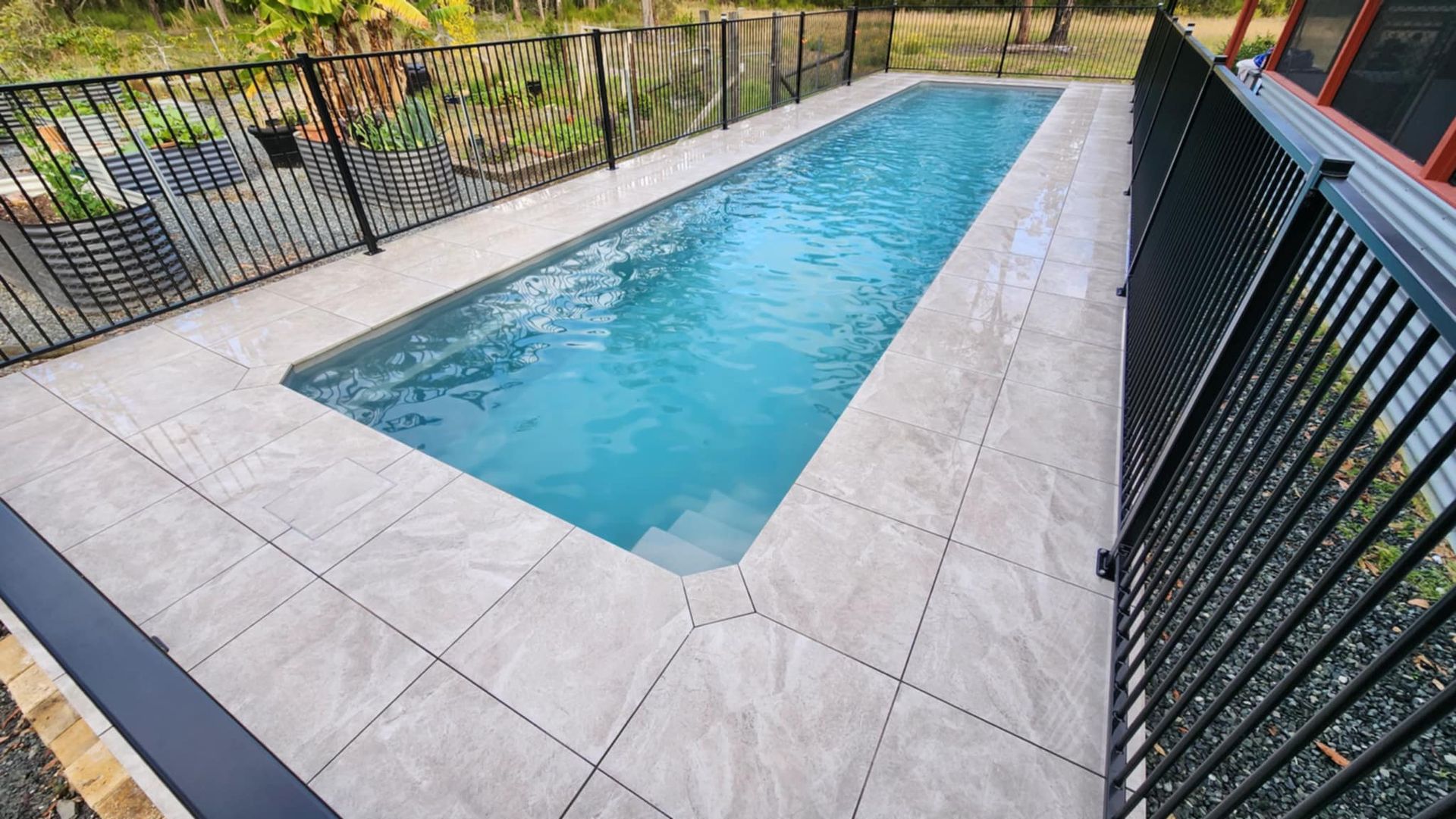 Rectangular pool with light-coloured tile surround, surrounded by a black fence and greenery. — Clearwater Pools Hallidays Point In Hallidays Point, NSW