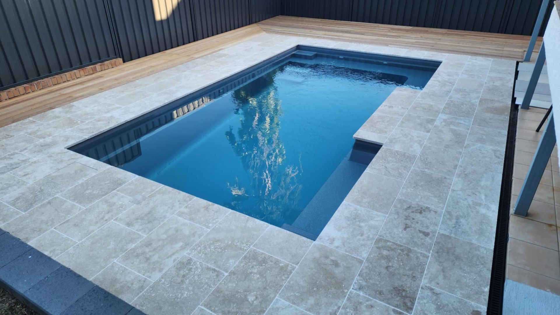 Rectangular in-ground pool surrounded by light stone tiles and wooden deck, set against a dark fence. — Clearwater Pools Hallidays Point In Hallidays Point, NSW