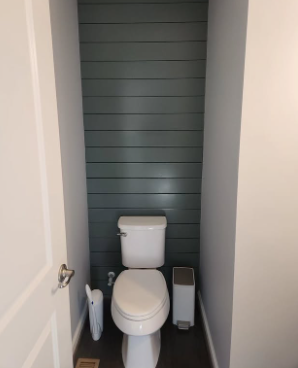 Toilet in a small bathroom with blue-green shiplap accent wall. Grey walls, white door and toilet. Trash can and toilet brush visible.