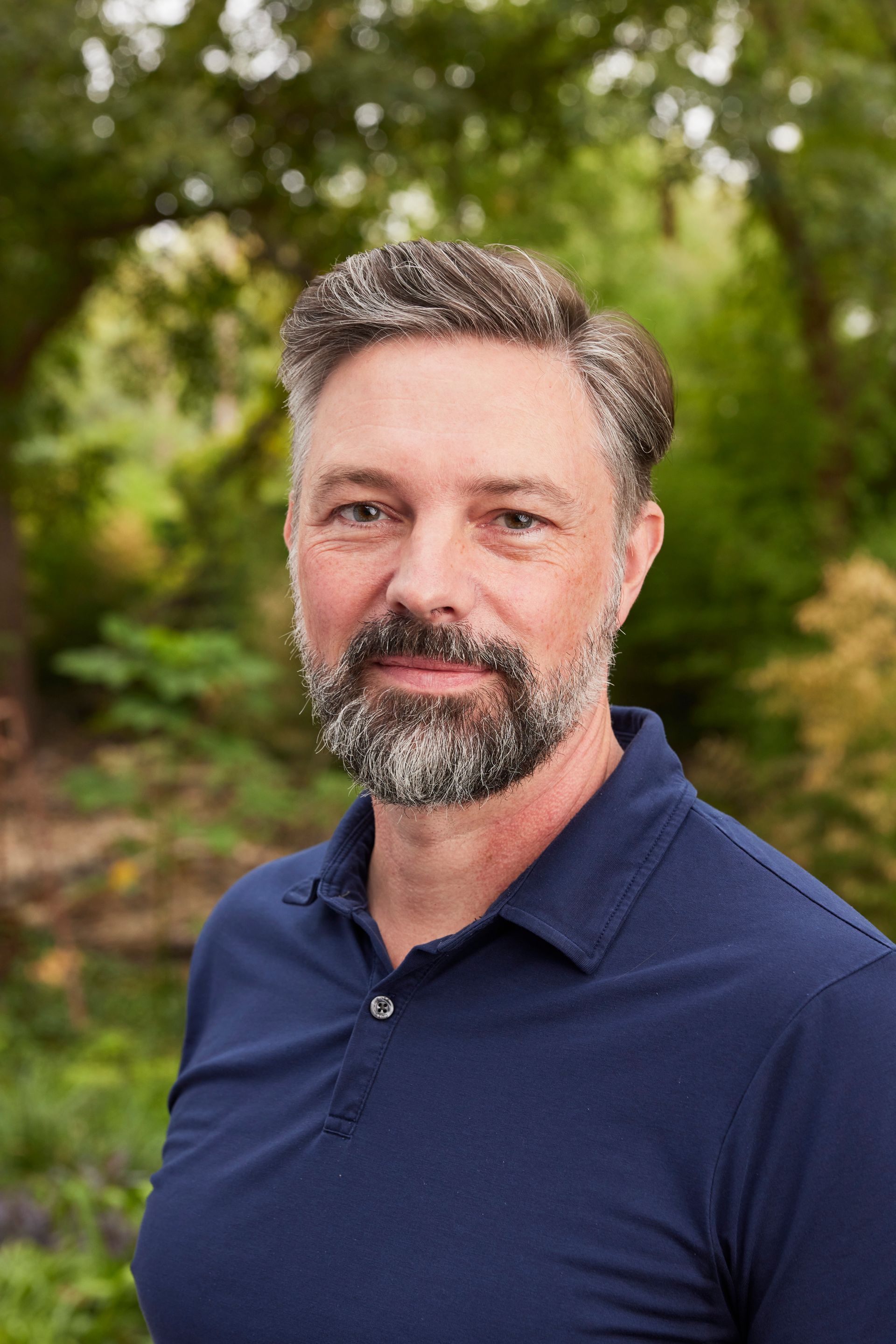 A man with a beard is wearing a blue polo shirt and standing in front of trees.