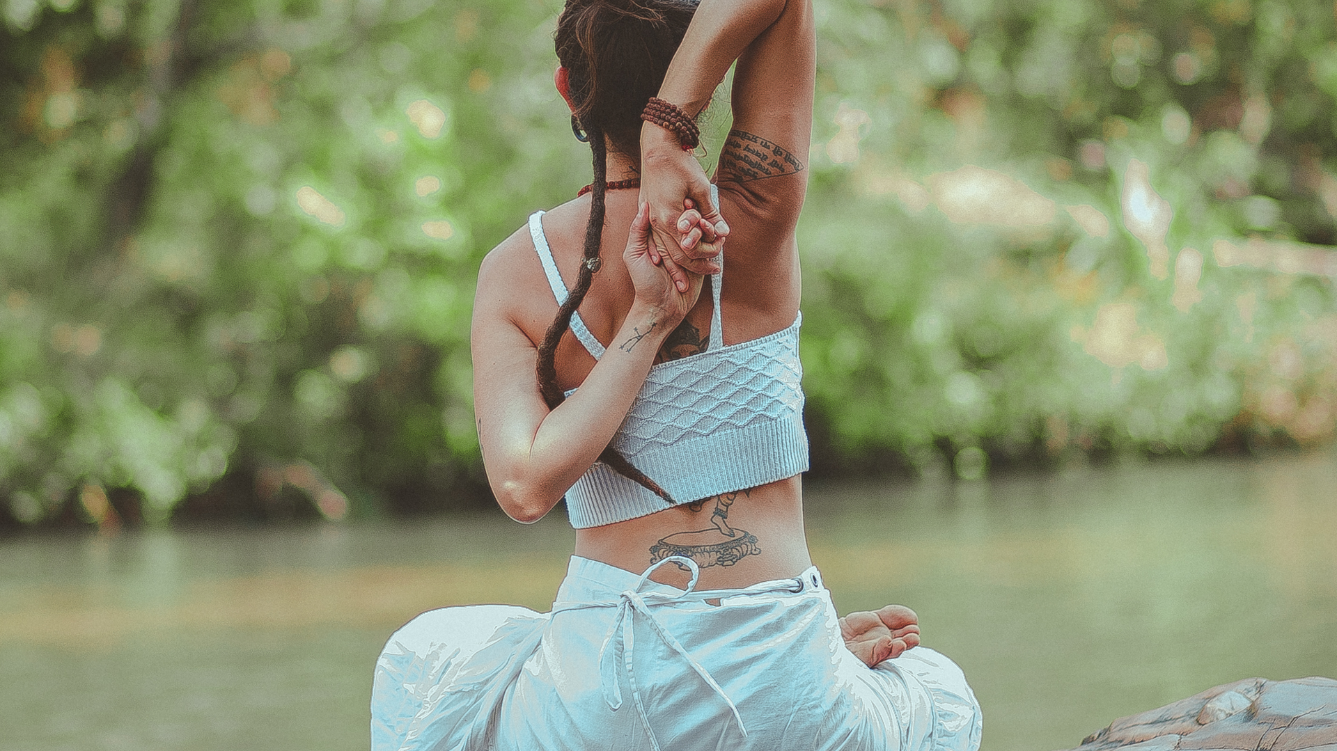 A woman is sitting on a rock by the water doing yoga.