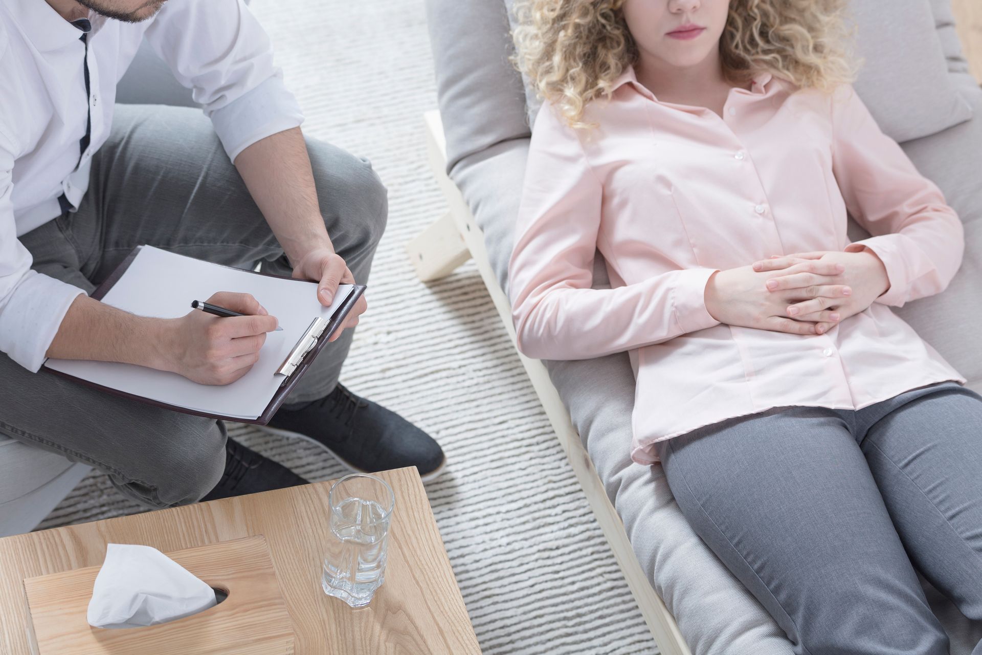 A woman is laying on a couch while a man writes on a clipboard.