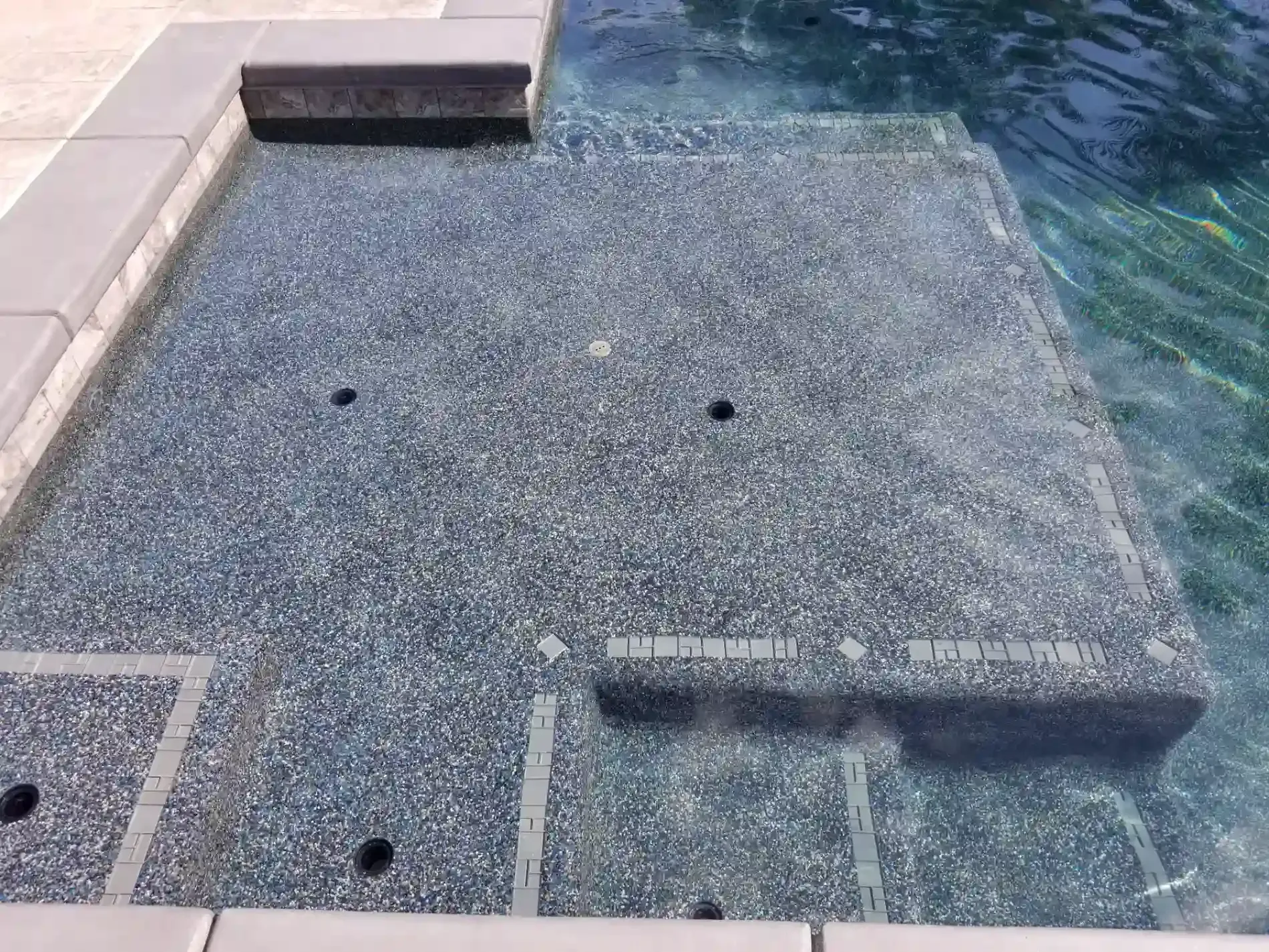 Poolside textured concrete deck beside blue water with steps and drain covers