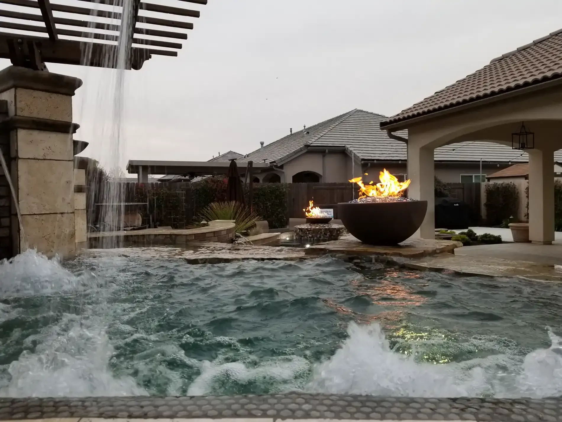 Backyard pool with rippling water and a fire bowl beside a covered patio at dusk