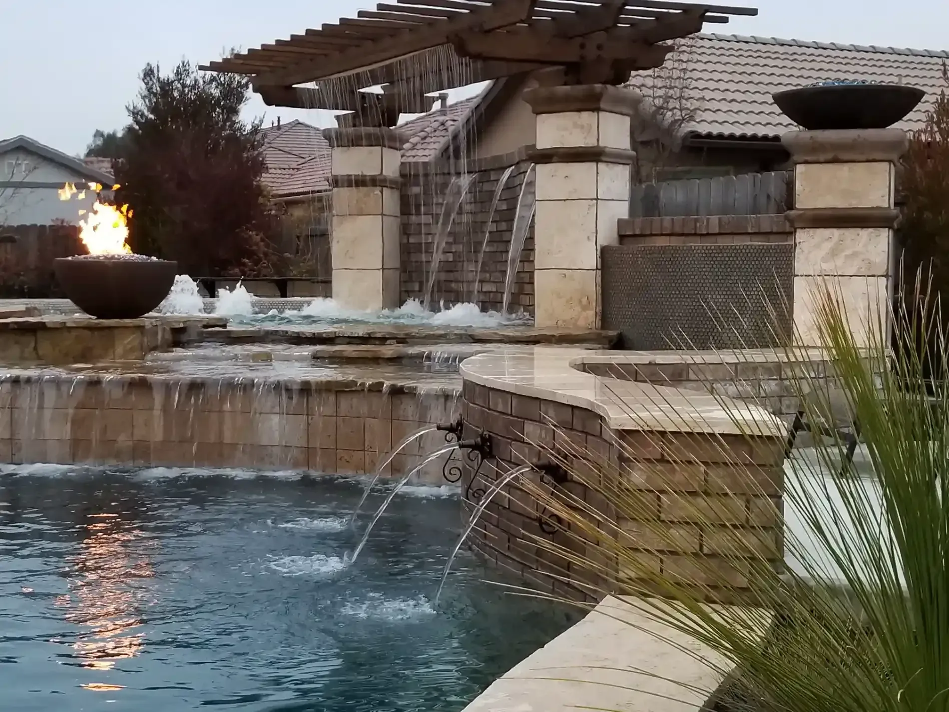 Backyard pool with a stone water feature, pergola, and lit fire bowl at dusk