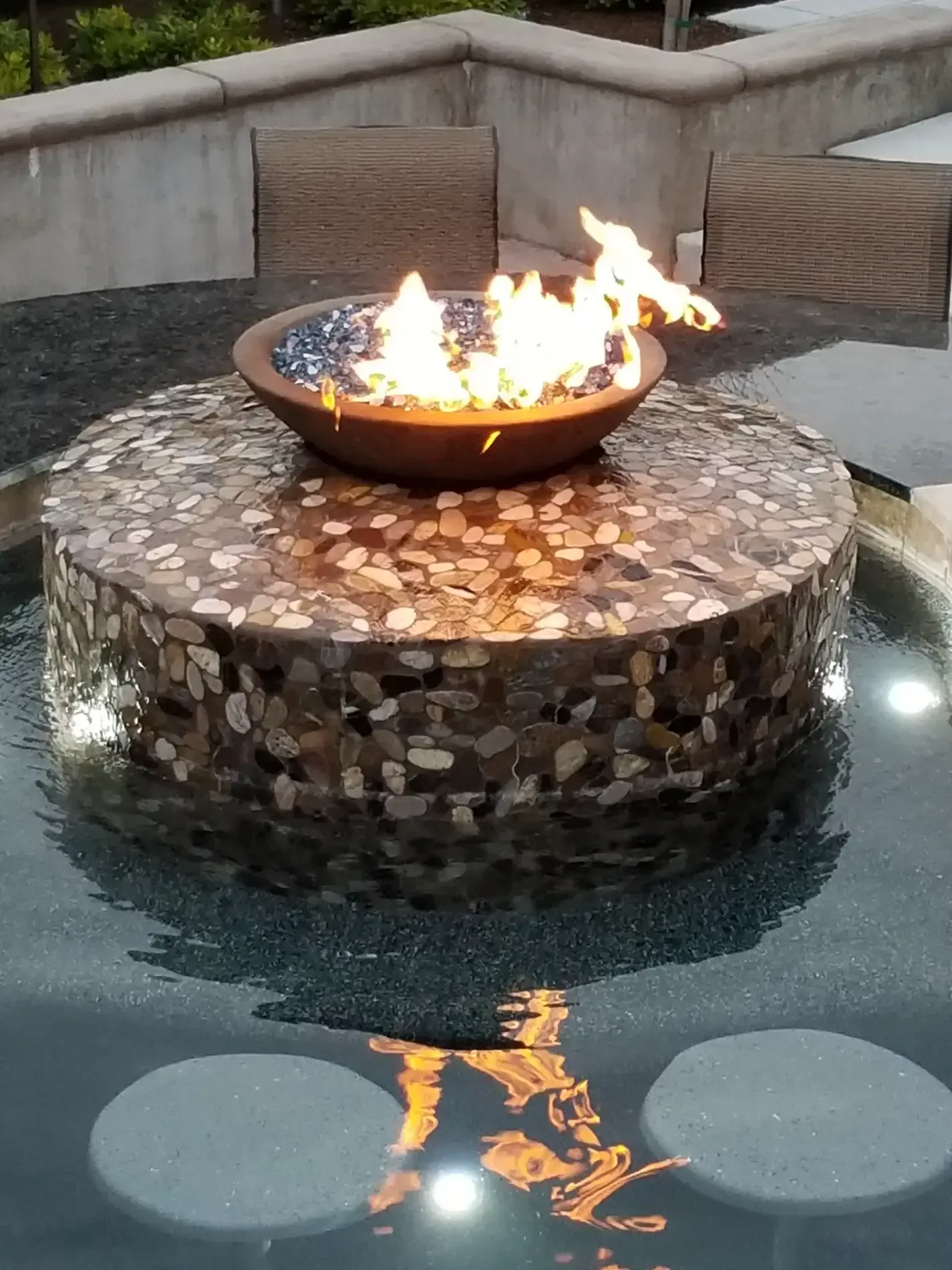 Round fire bowl on stone pedestal reflecting in a shallow water feature at a modern patio garden
