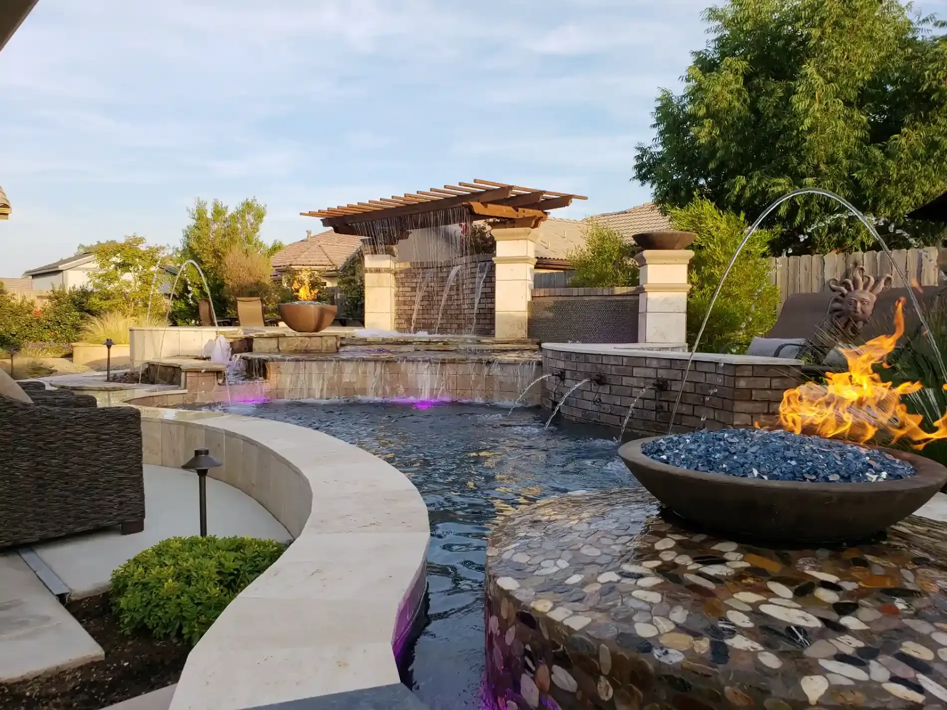 Modern backyard water feature with stone patio, pergola, fire bowl, and purple-lit waterfall