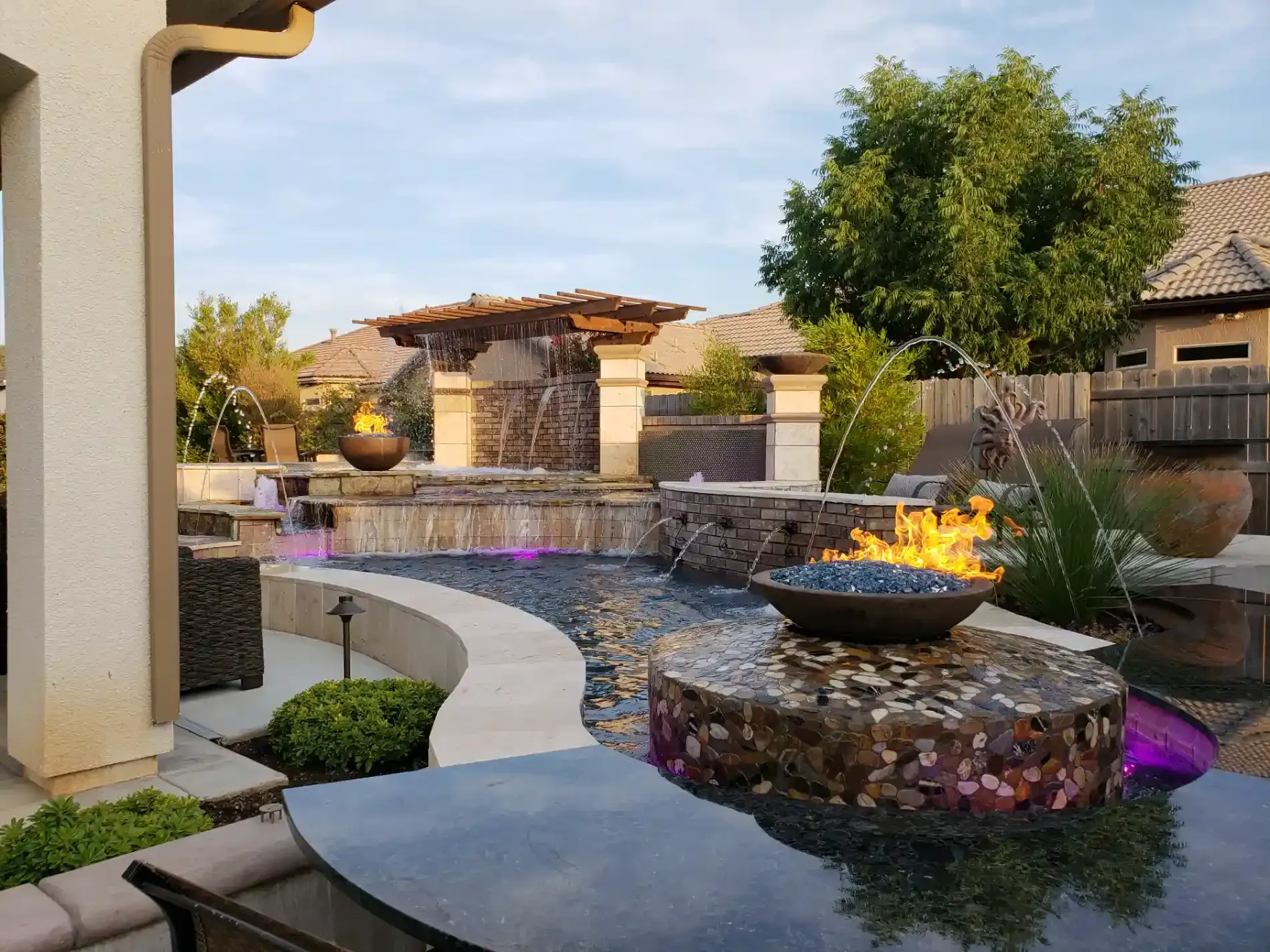 Modern backyard patio with curved stone paths, waterfall pool, and glowing fire bowl at dusk