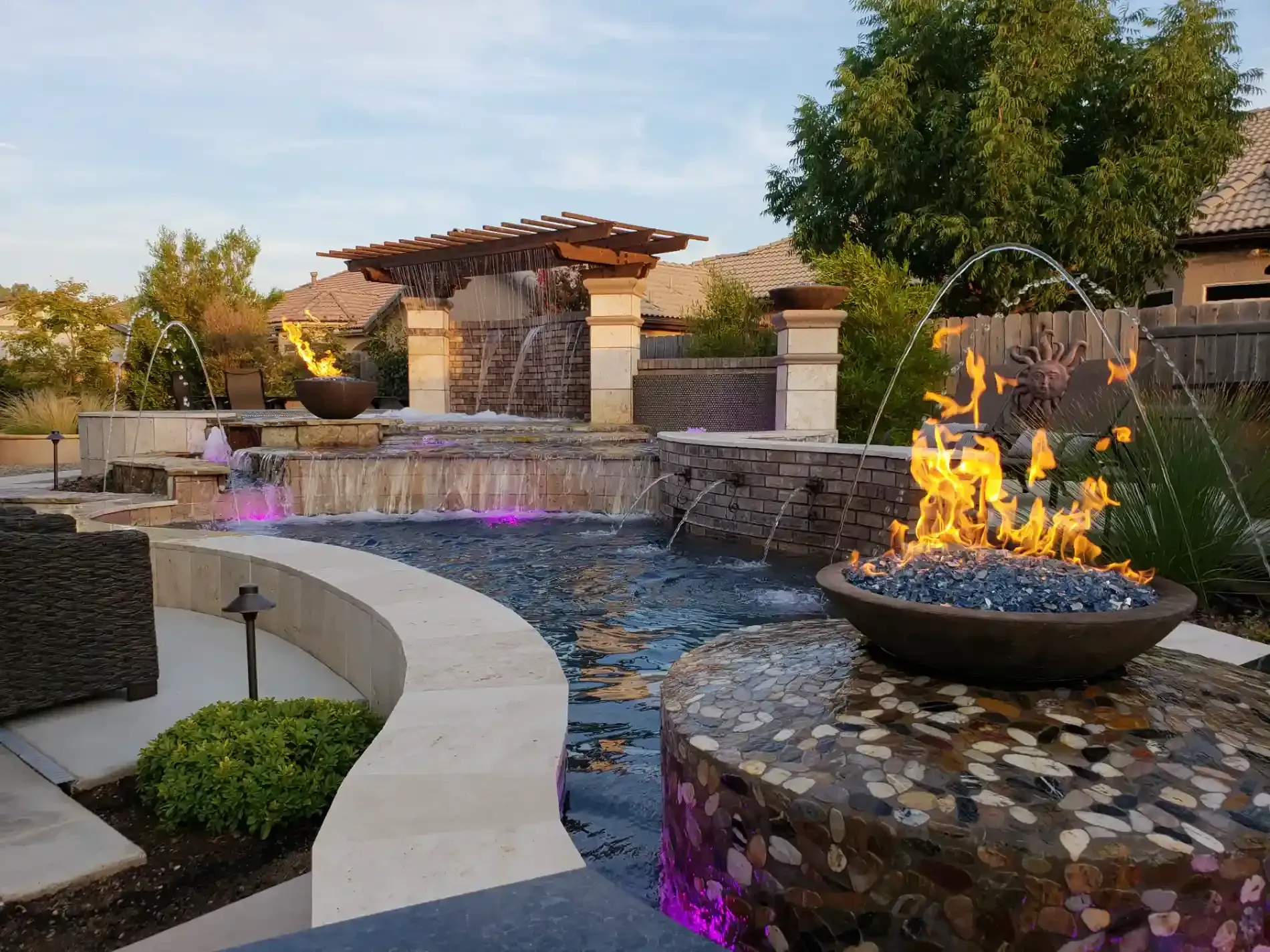 Modern backyard water feature with stone walls, flowing water, and two fire bowls at dusk