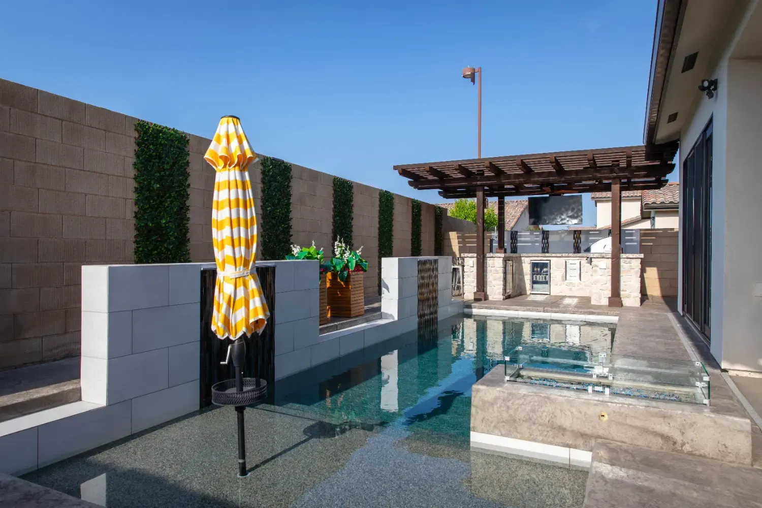 Modern backyard patio with a swimming pool, pergola, and yellow striped umbrella under a clear blue sky