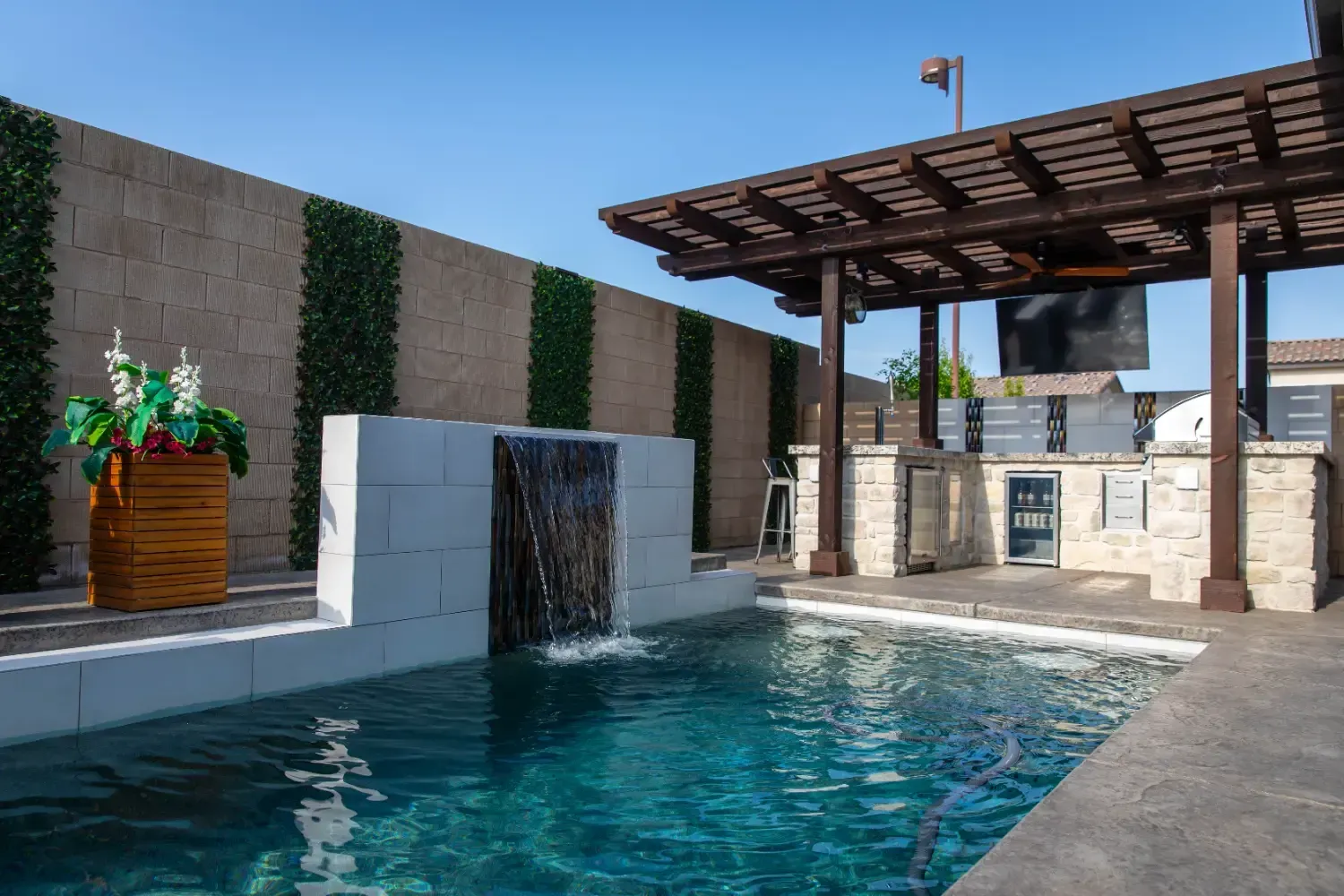 Modern patio pool with a waterfall feature, pergola shade, and stone walls under a clear blue sky