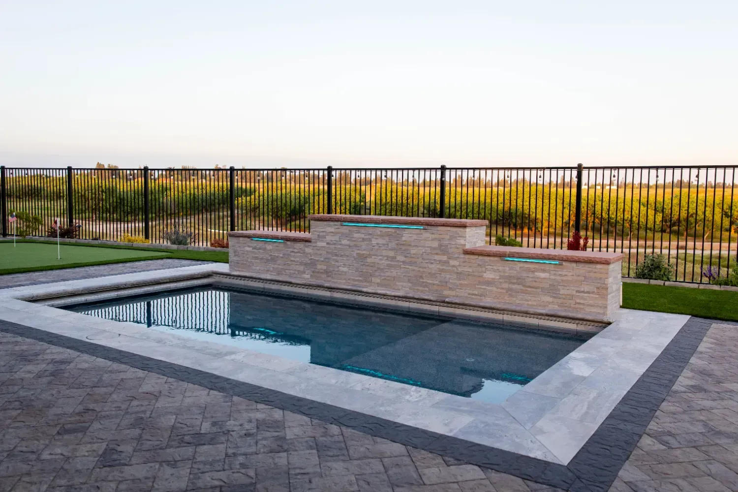 Rectangular backyard pool with stone decking, black fence, and landscaped yard at sunset