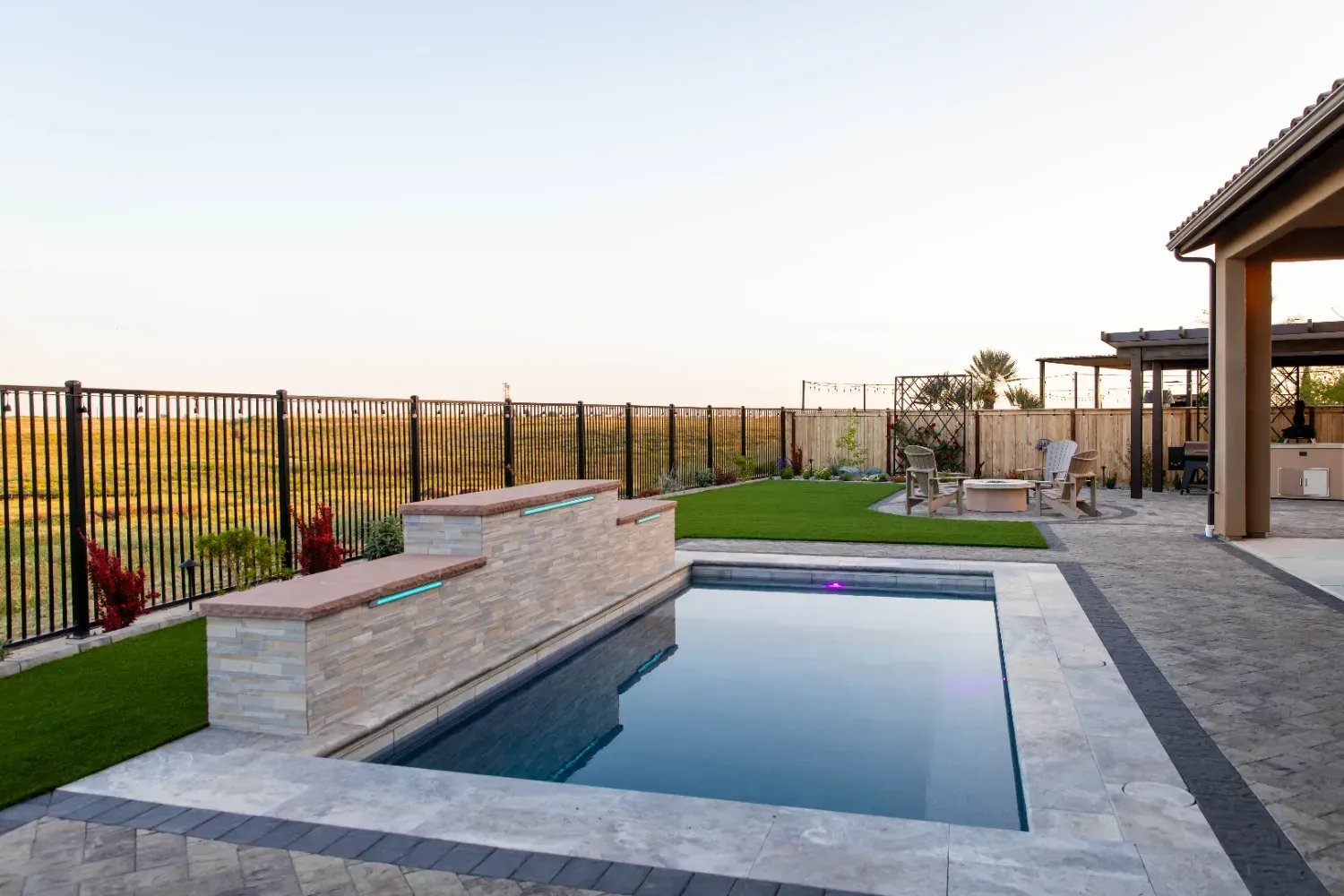 Modern backyard with a rectangular pool, stone deck, green lawn, and metal fence at sunset