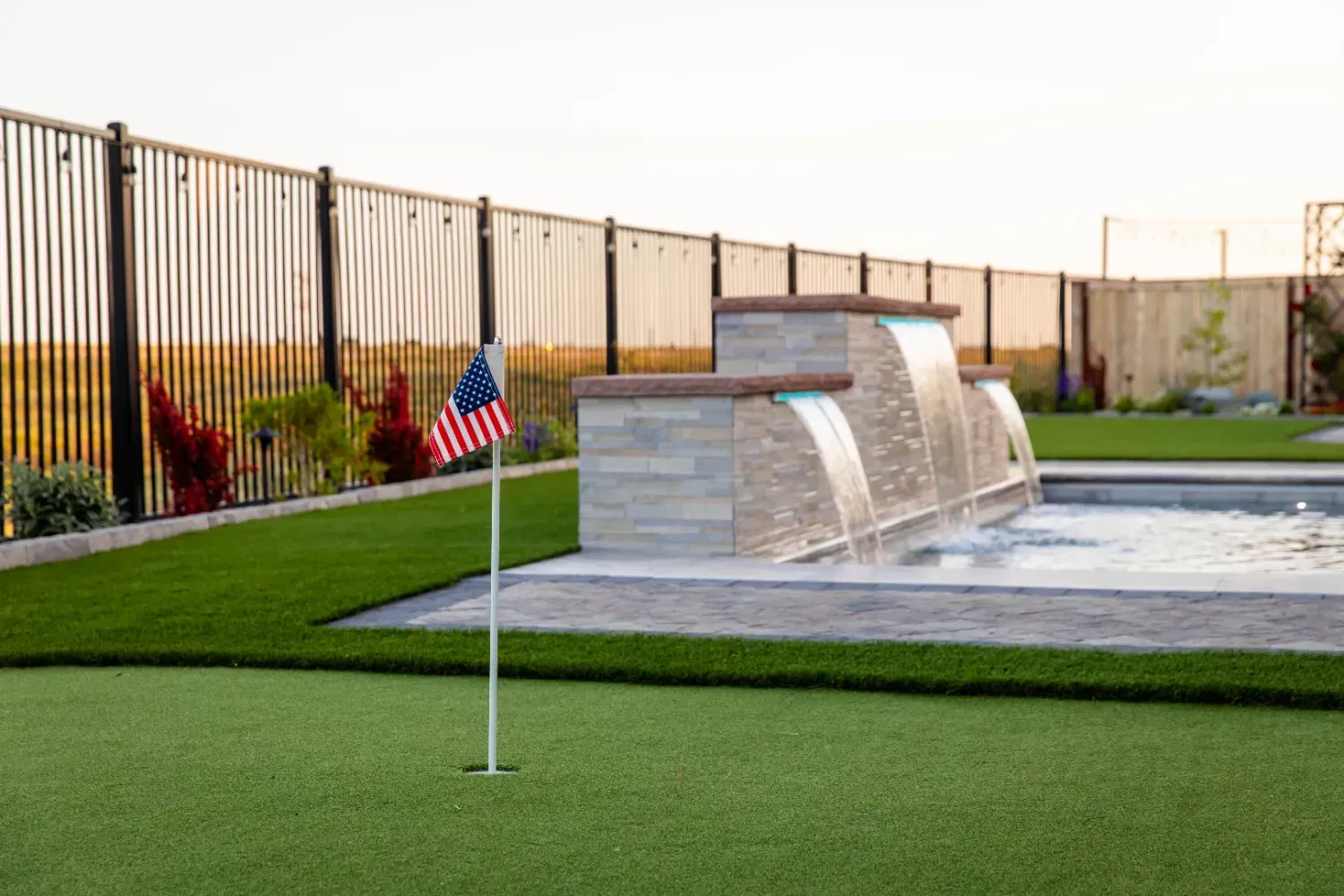 Modern memorial fountain beside a lawn, with a small U.S. flag in the foreground and a metal fence behind