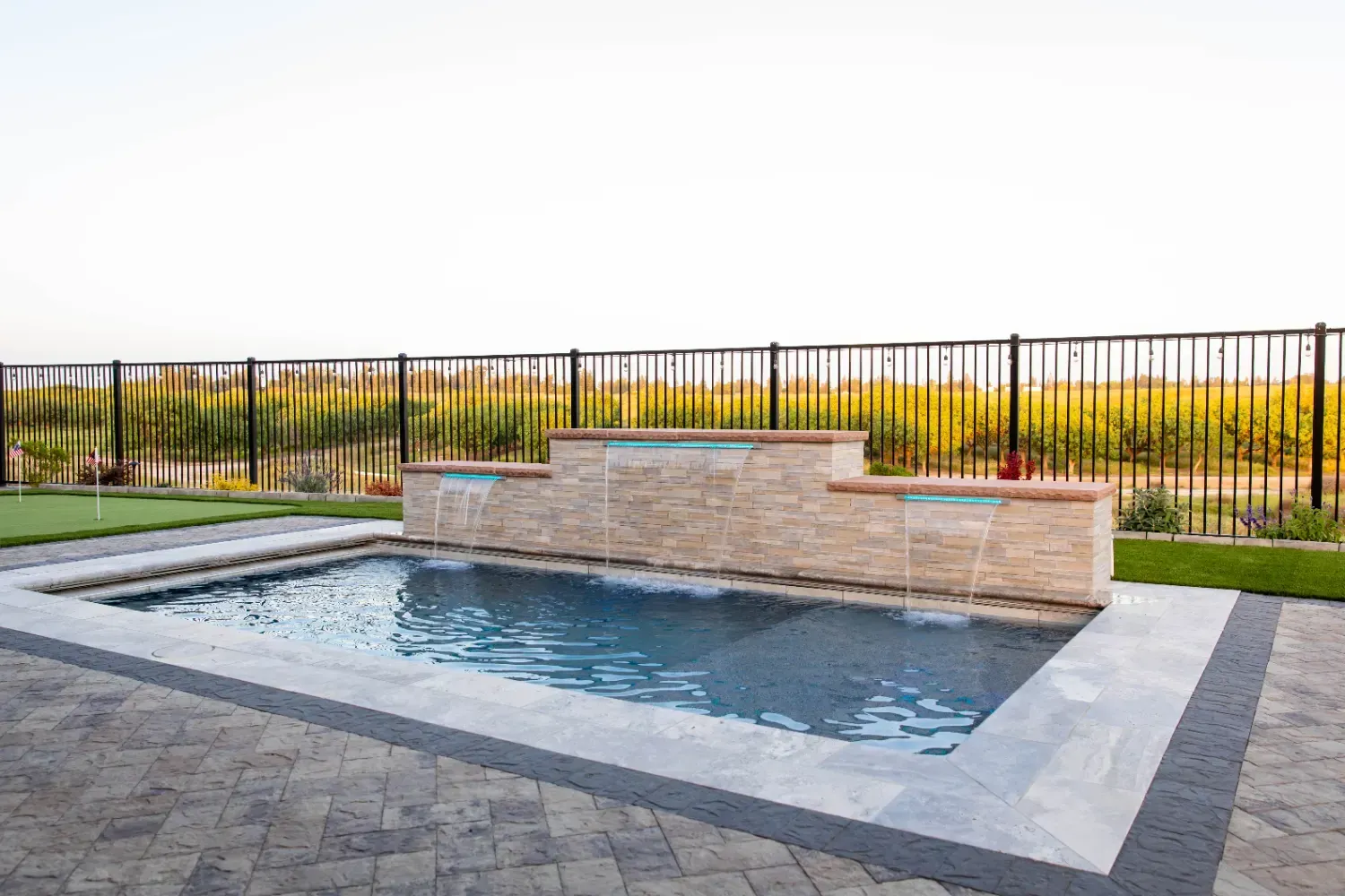 Modern backyard pool with a stone water feature and black metal fence overlooking a grassy field