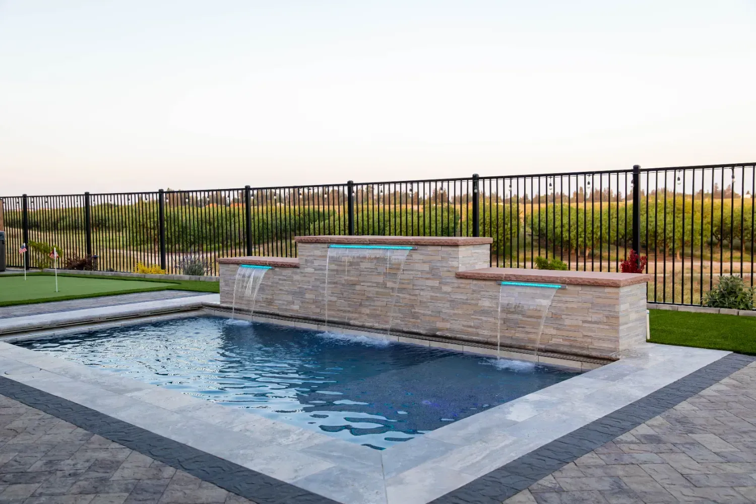 Rectangular backyard pool with a stone water feature and metal fence at sunset