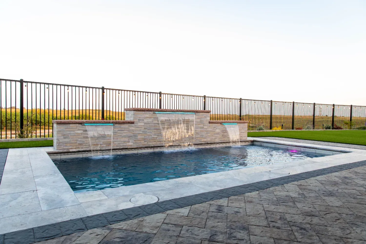 Rectangular stone fountain pool with cascading water, set in a landscaped outdoor patio with fencing and hills beyond