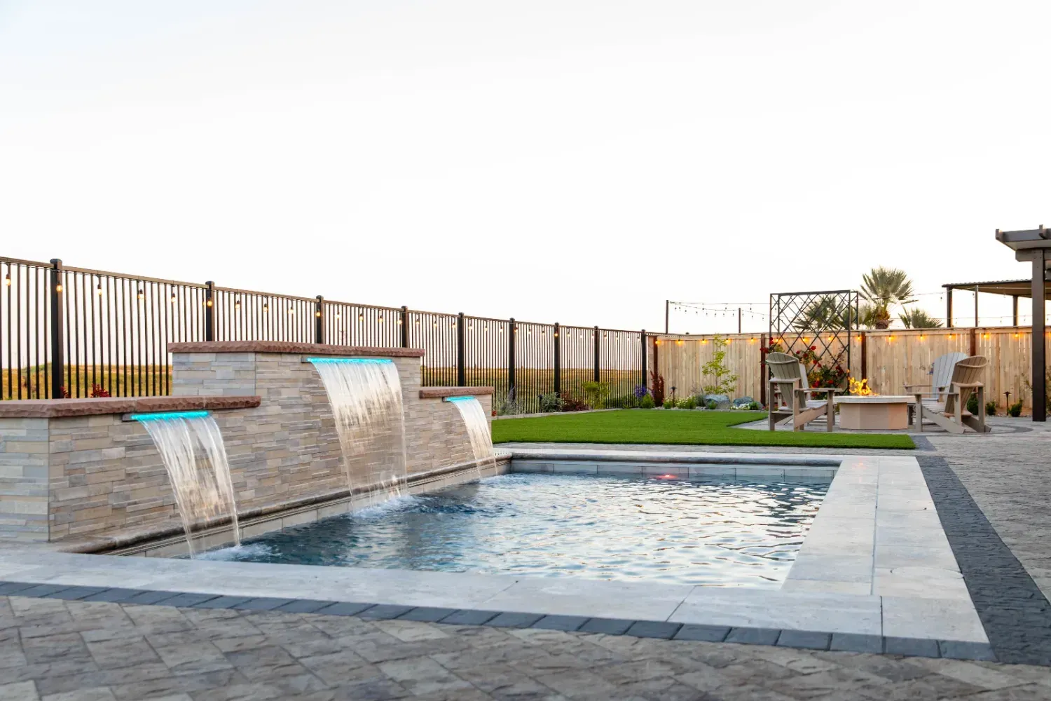 Modern backyard pool with waterfall feature, stone decking, and fenced patio at sunset
