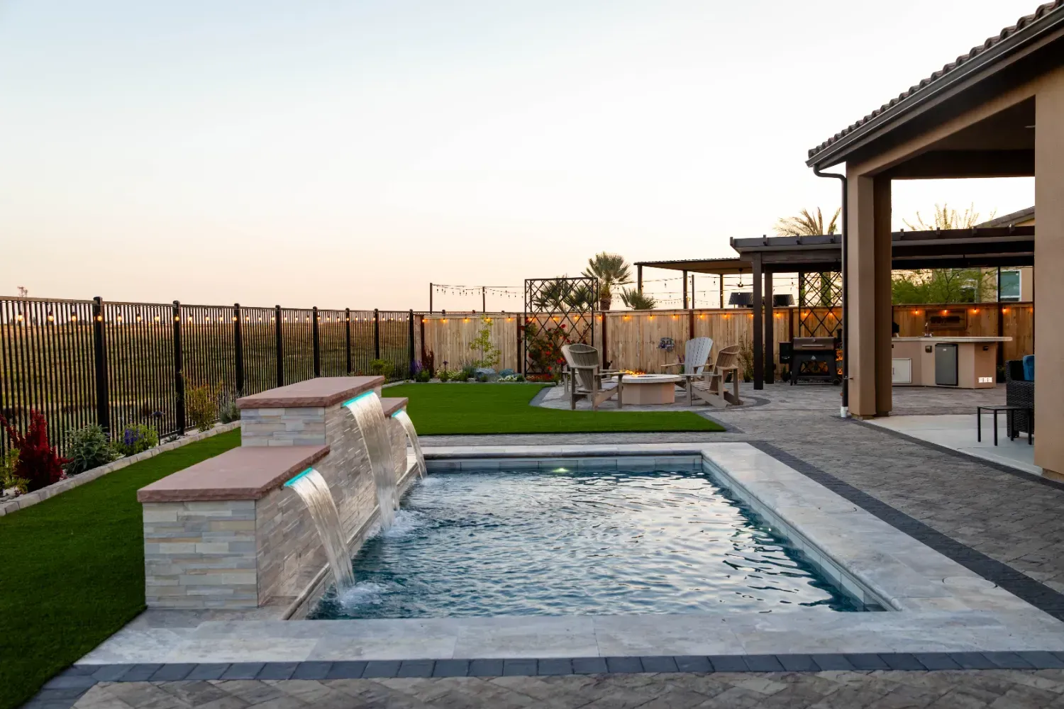 Modern backyard with a small pool, stone lounge ledge, patio seating, and a covered outdoor area at sunset