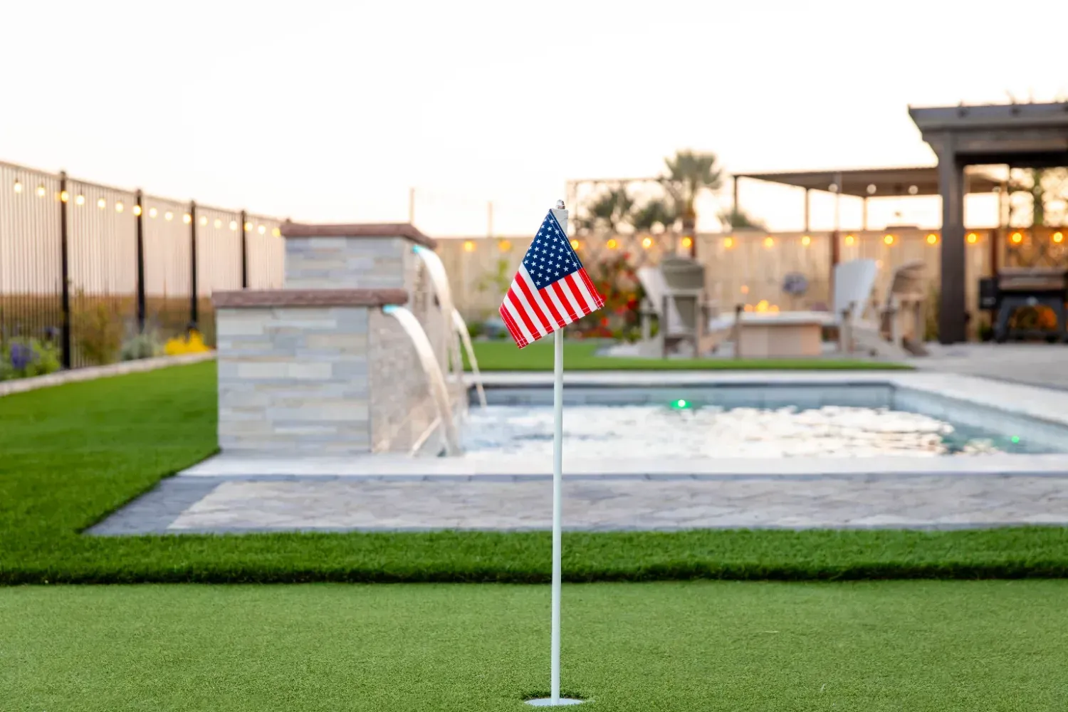 Small American flag on a pole beside a backyard pool and green lawn at sunset