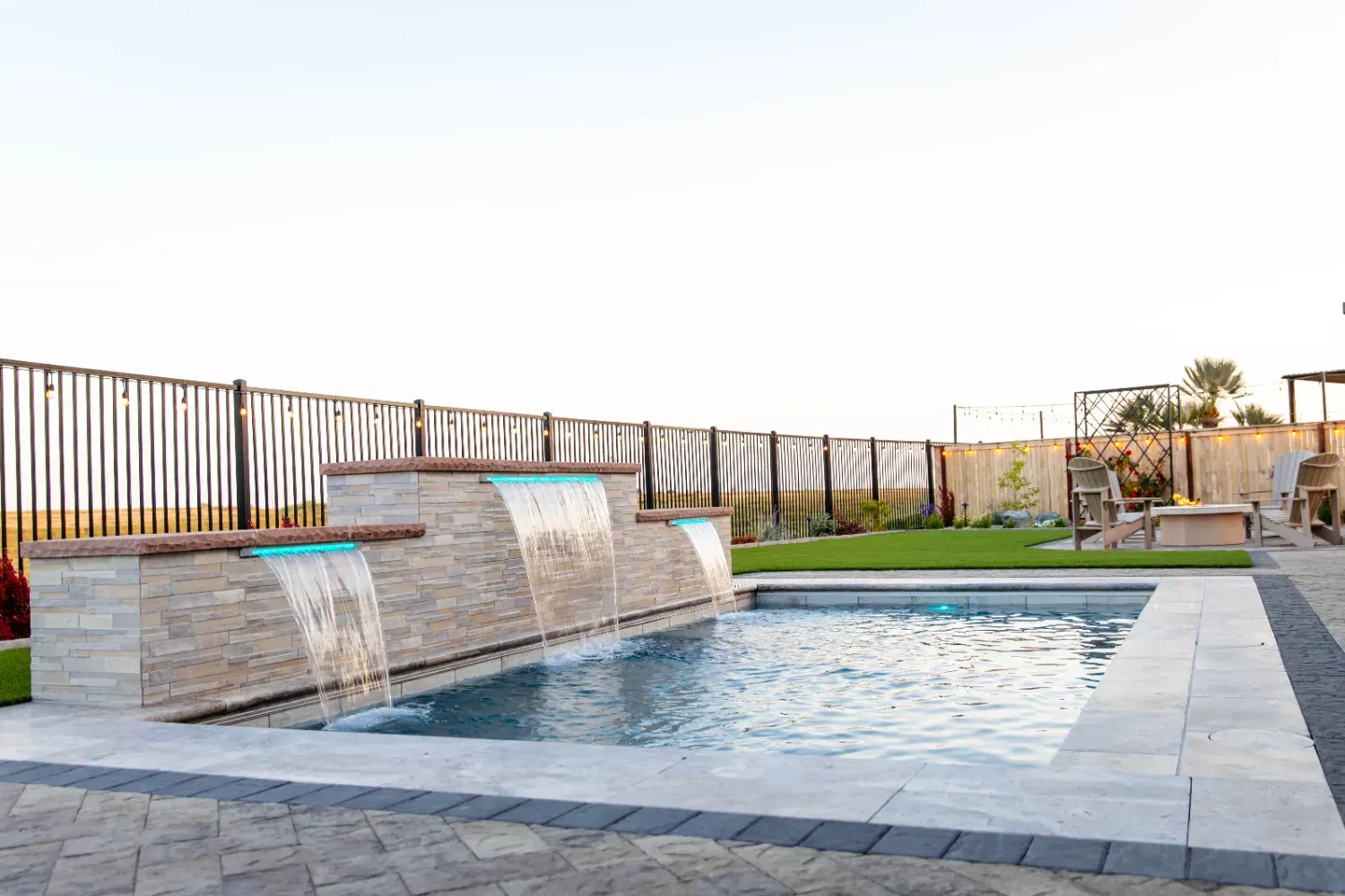 Modern backyard pool with stone waterfall, tiled deck, and grassy lawn at sunset