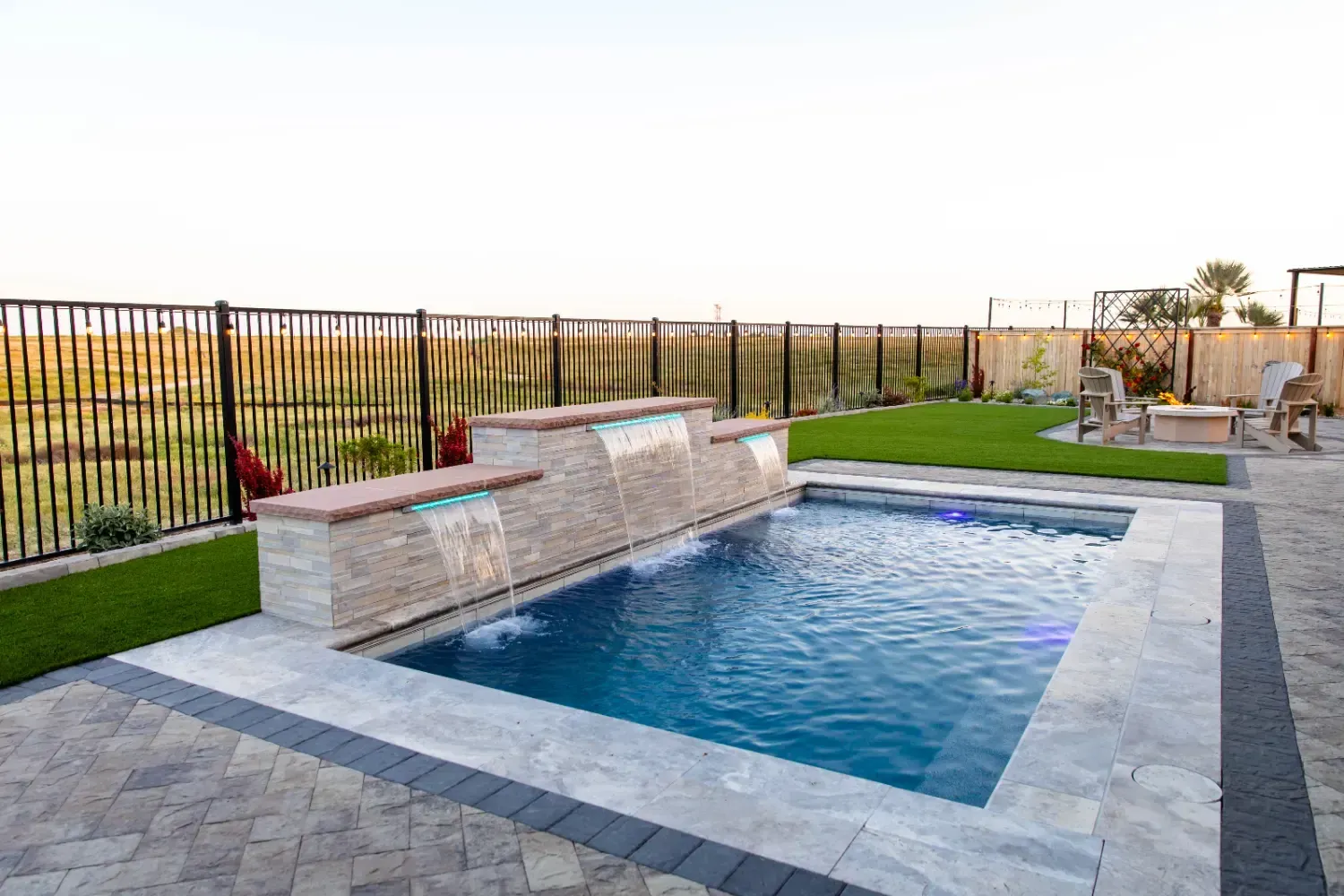 Modern backyard pool with stone deck, waterfall wall, and fenced lawn area