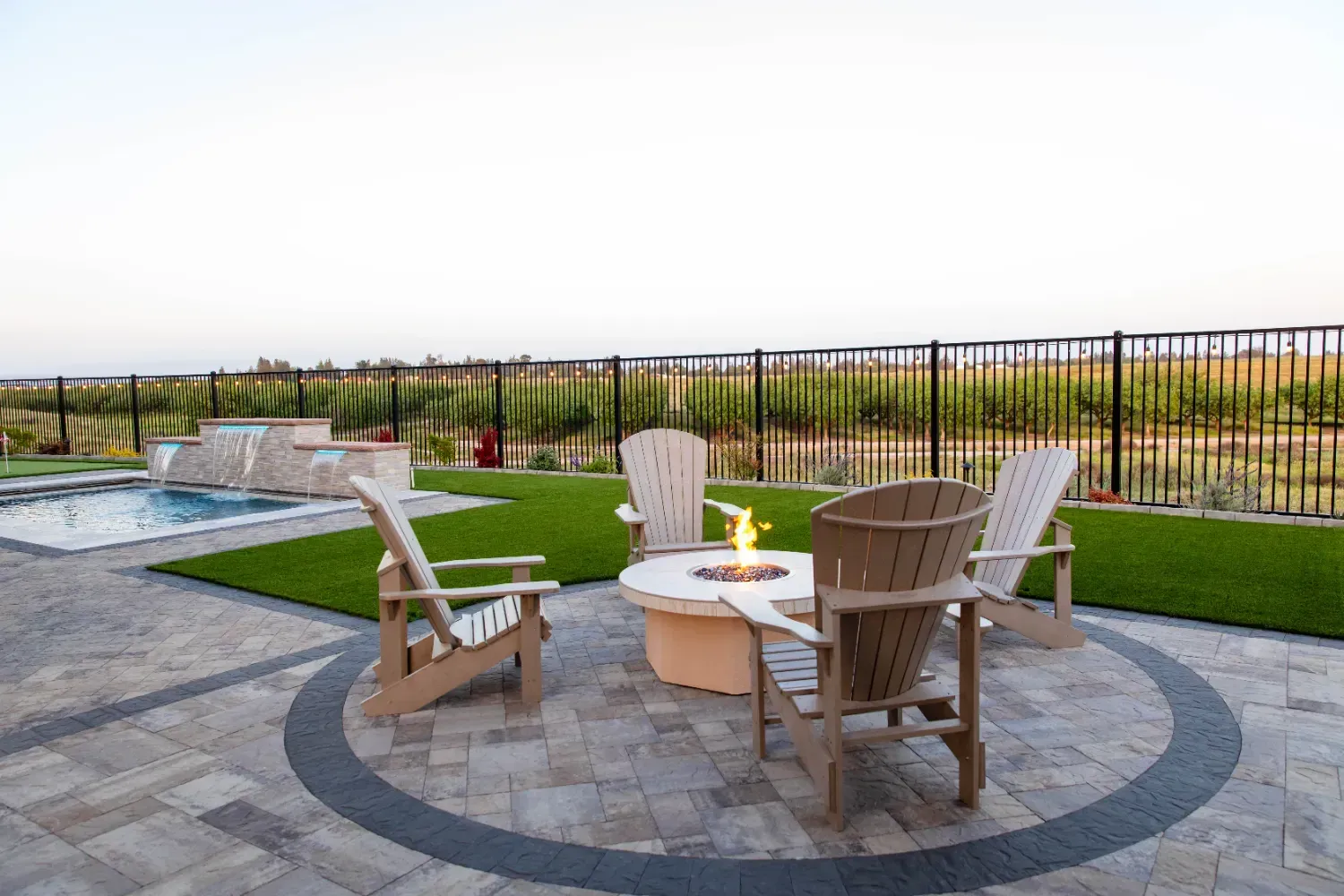 Outdoor patio with circular stone fire pit, four wooden chairs, and a fenced lawn at sunset
