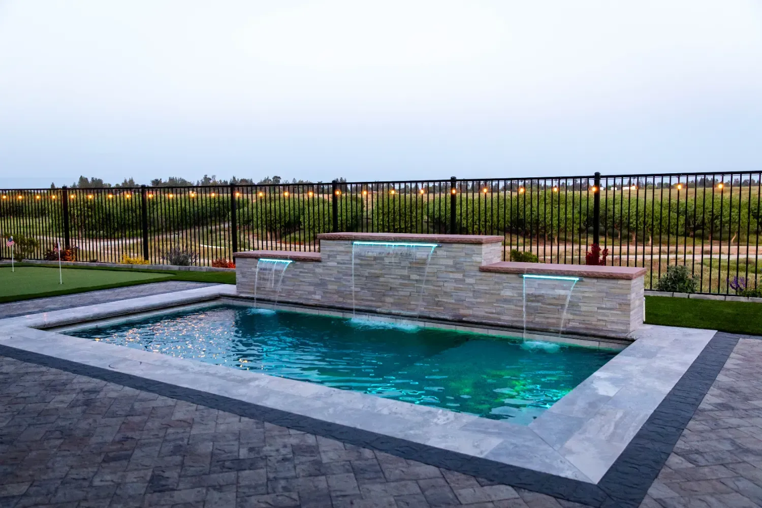 Small backyard swimming pool with stone decking, water feature wall, and black metal fence at dusk