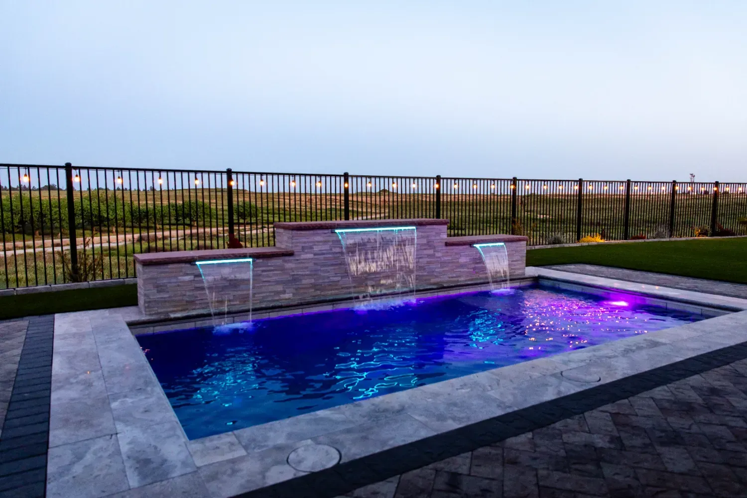 Modern backyard pool with blue and purple LED lighting and a stone waterfall feature at dusk