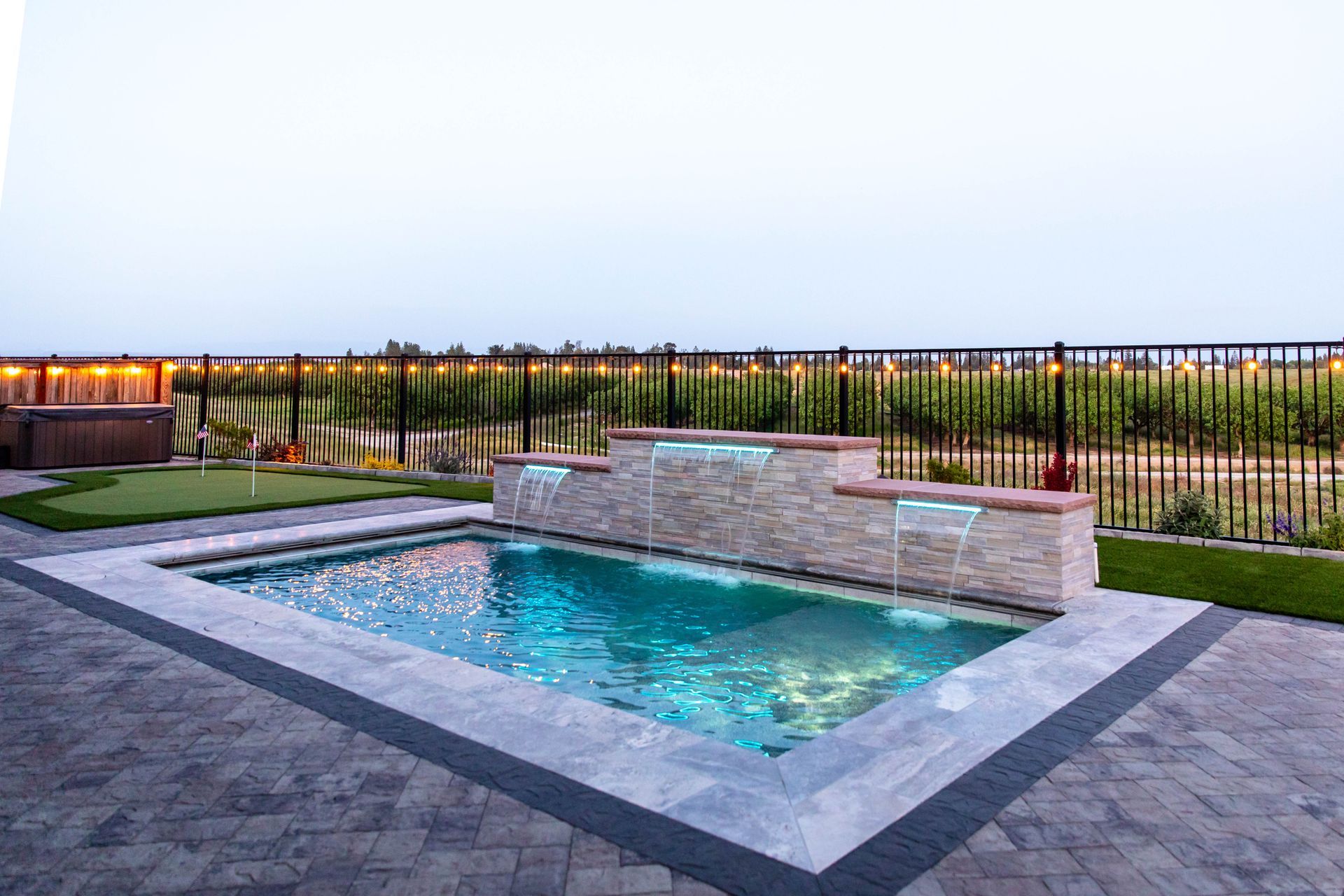 Modern backyard with a lit rectangular pool, stone patio, and fenced landscape at dusk