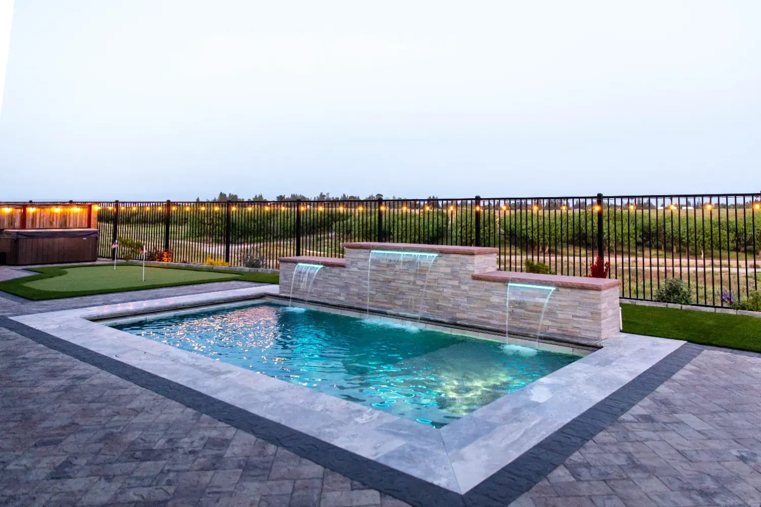 Modern backyard patio with a small rectangular pool, stone bench, and fenced lawn at dusk