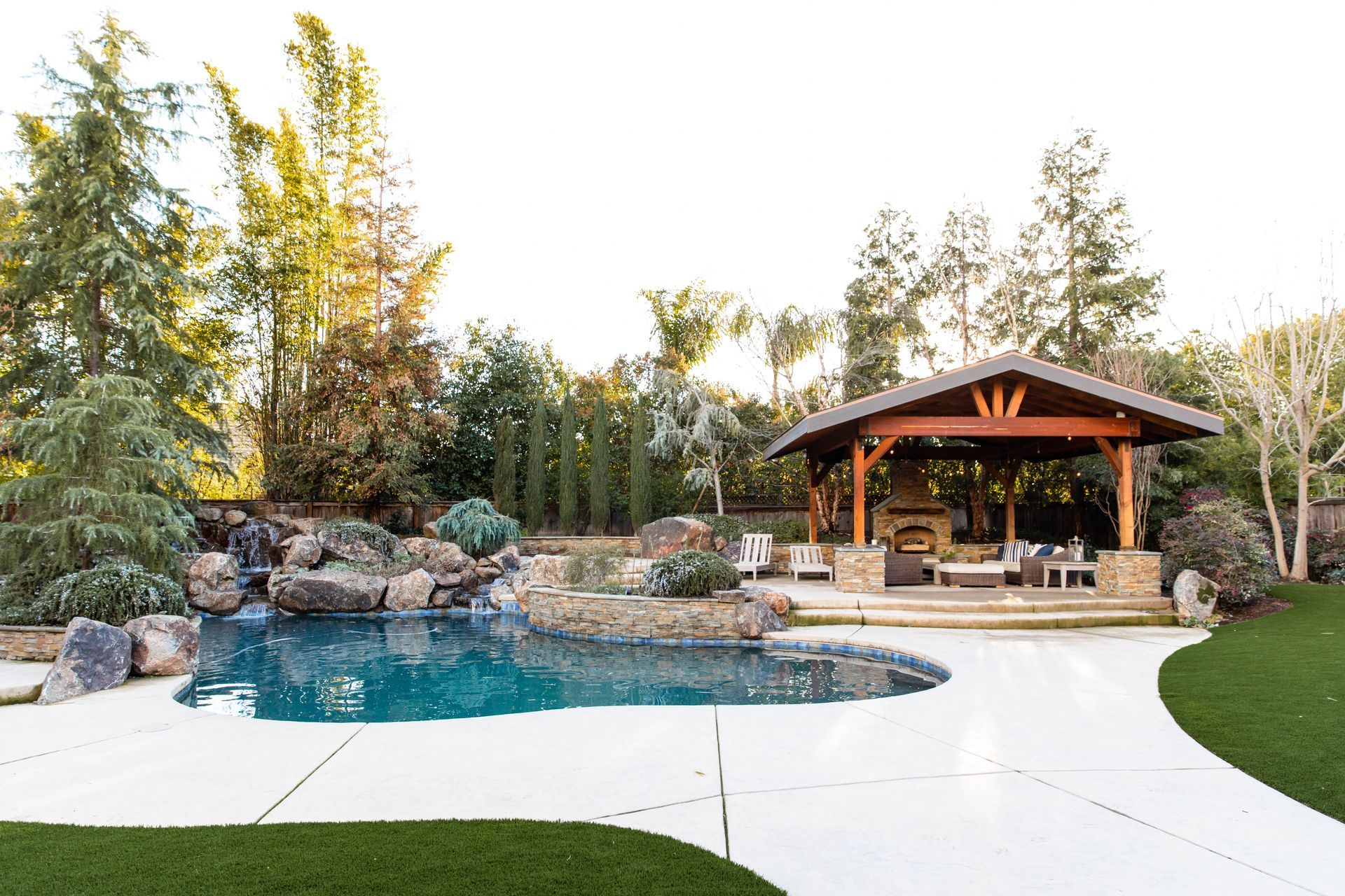 Backyard pool with waterfall rocks, white patio, and a wooden gazebo surrounded by trees
