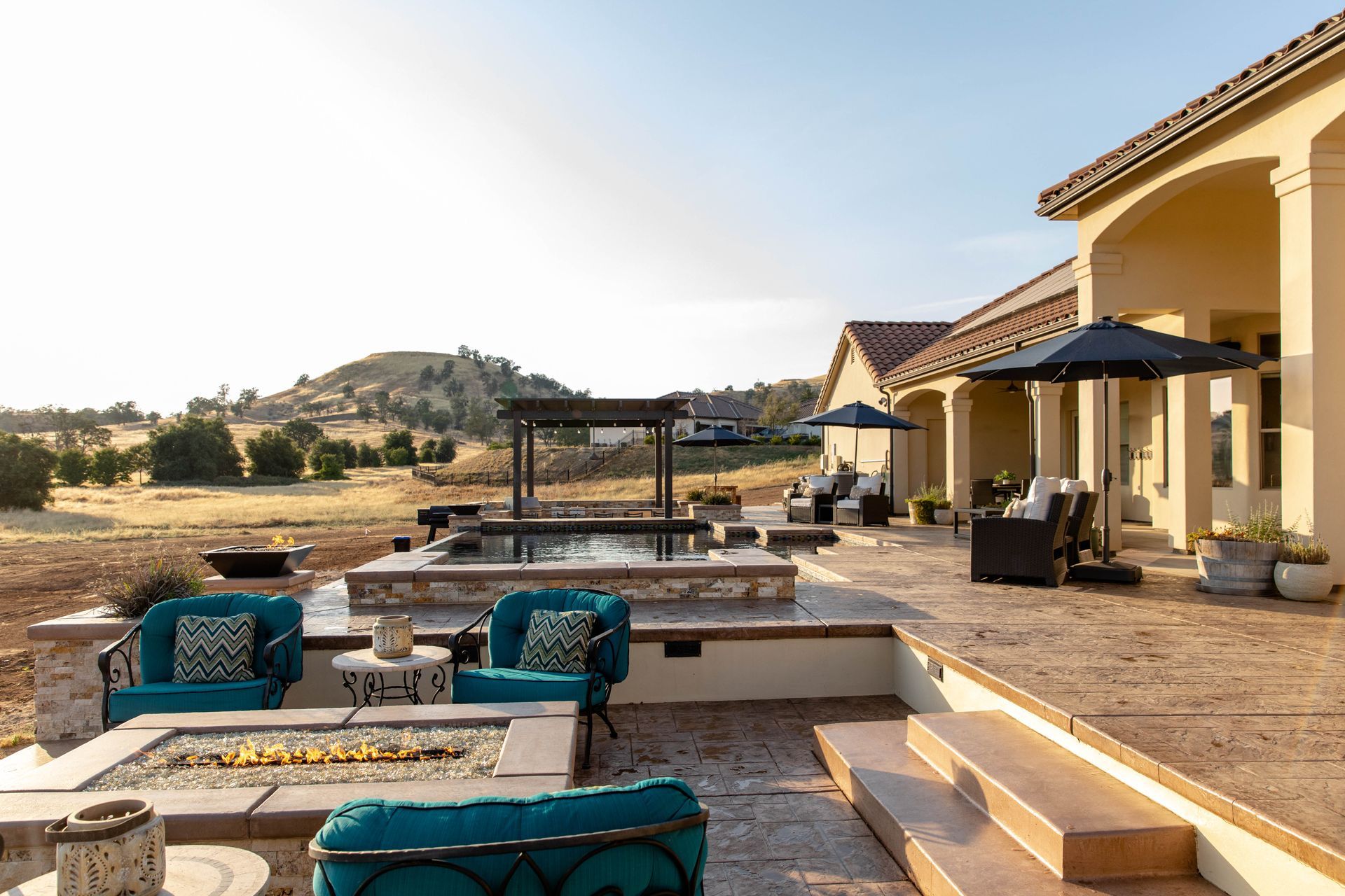 Modern patio with turquoise chairs, fire pit, and hillside view beside a beige house