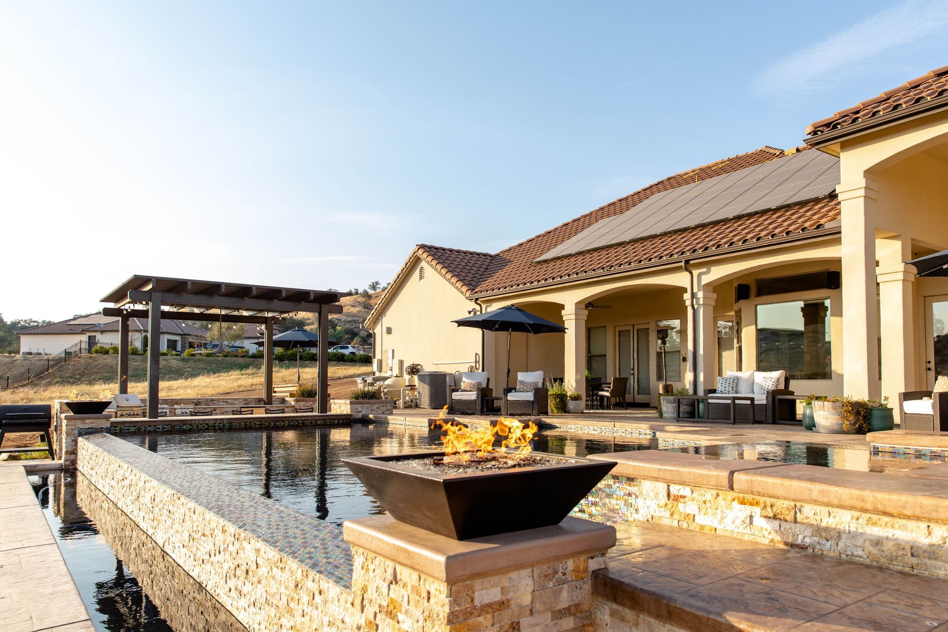 Luxury patio with stone fire pit, pool, and covered seating beside a stucco villa under a blue sky